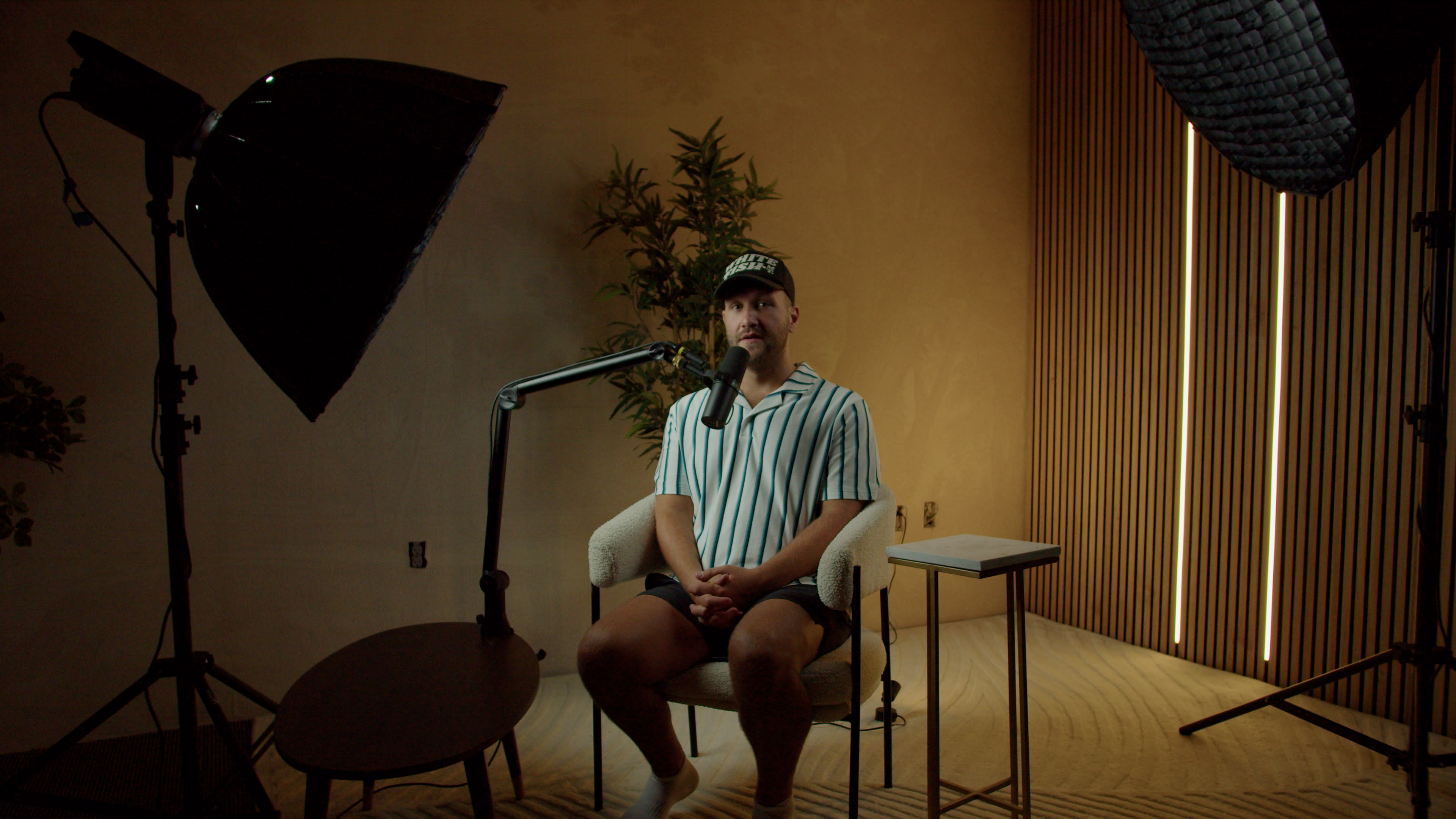 A man sits on a chair in a well-lit room with a plant and minimal furniture.