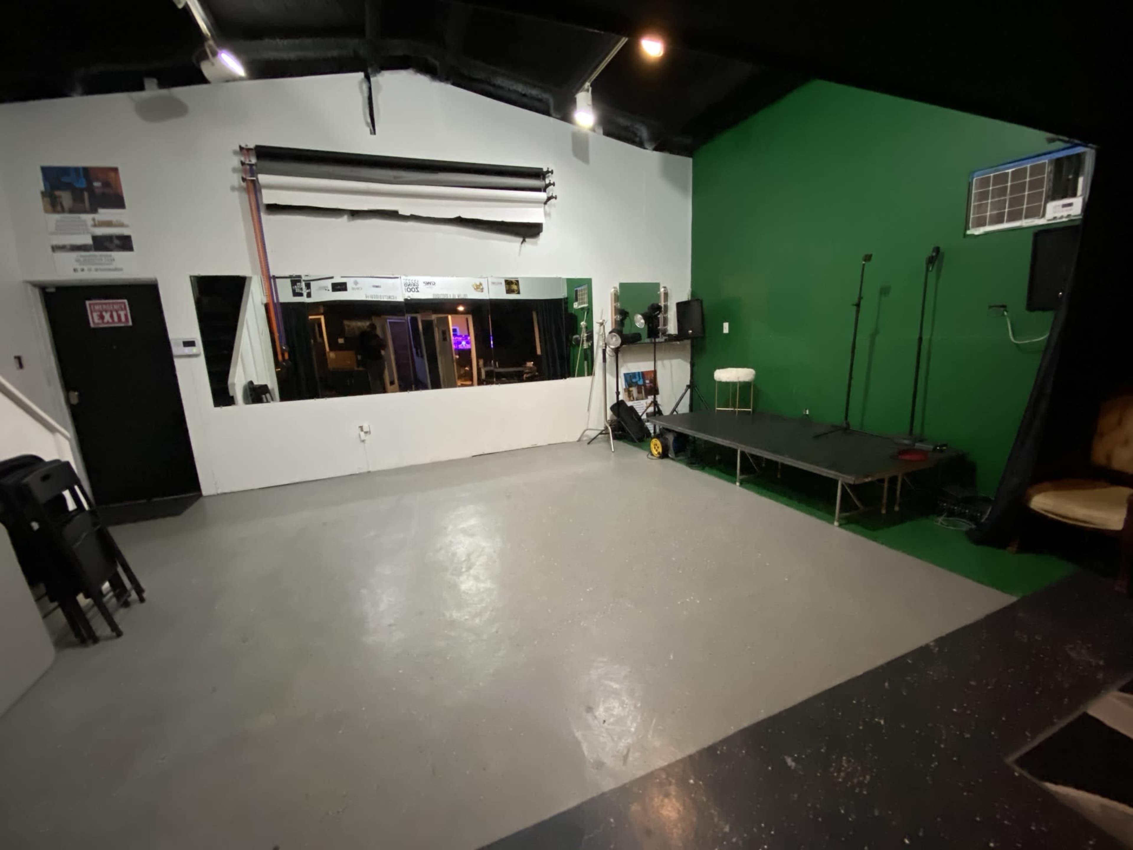 A performance space featuring a green wall, a small stage, audio equipment, and a large mirror opposite the stage.