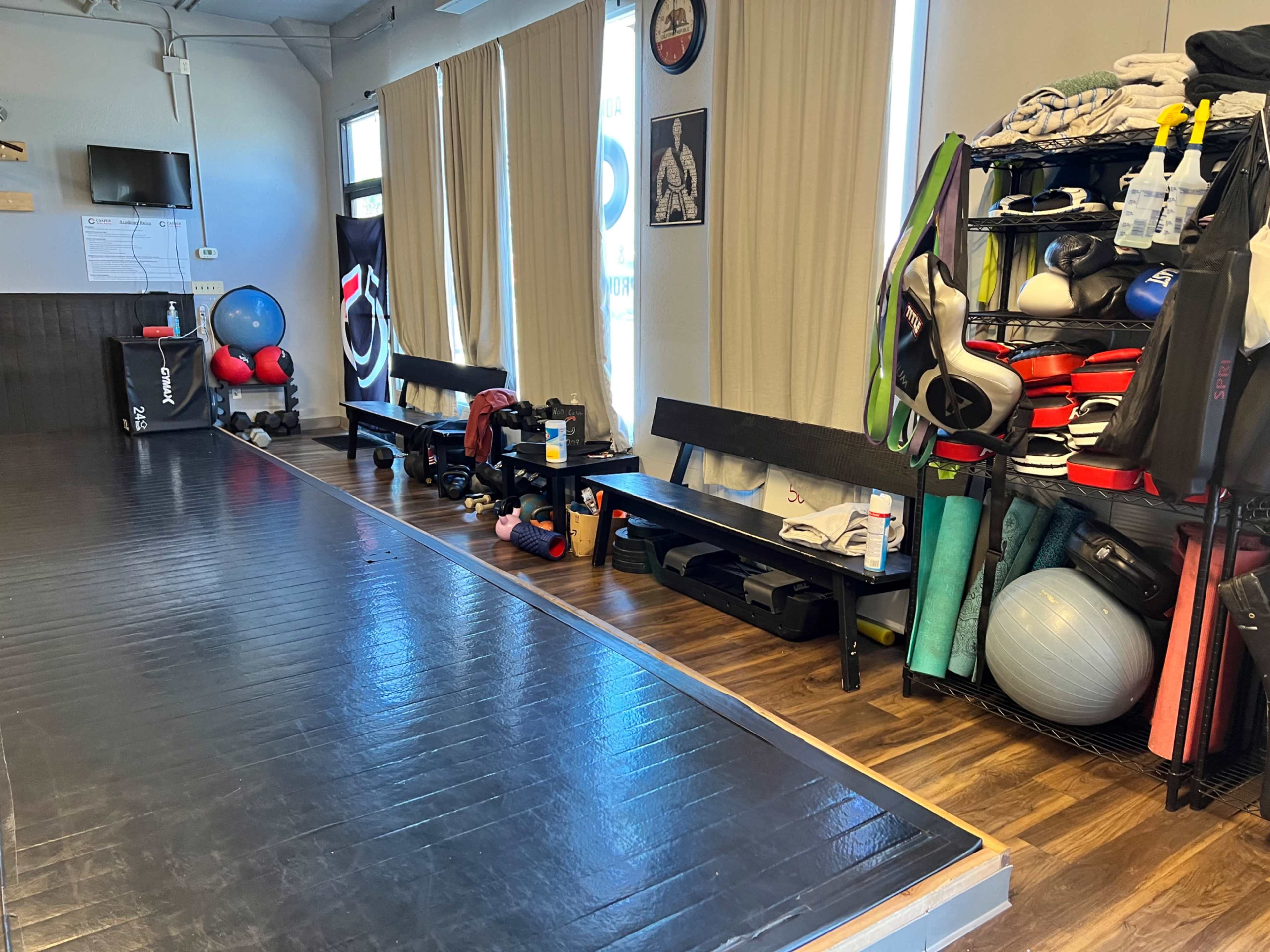 The image shows a fitness studio with a black mat area on the floor, benches along the side, and shelves stocked with various training equipment and exercise gear.