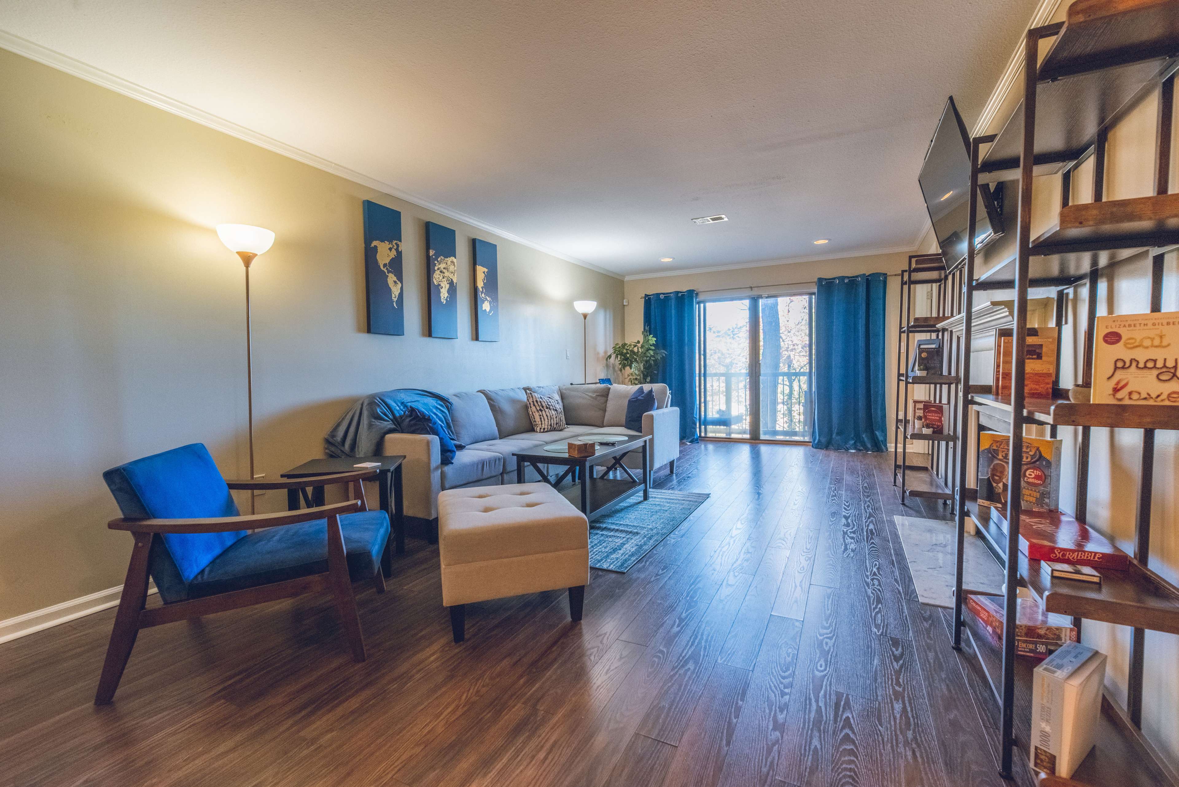 The living room features a couch, an armchair, a coffee table, and a large window with blue curtains, surrounded by bookshelves and decorative items.