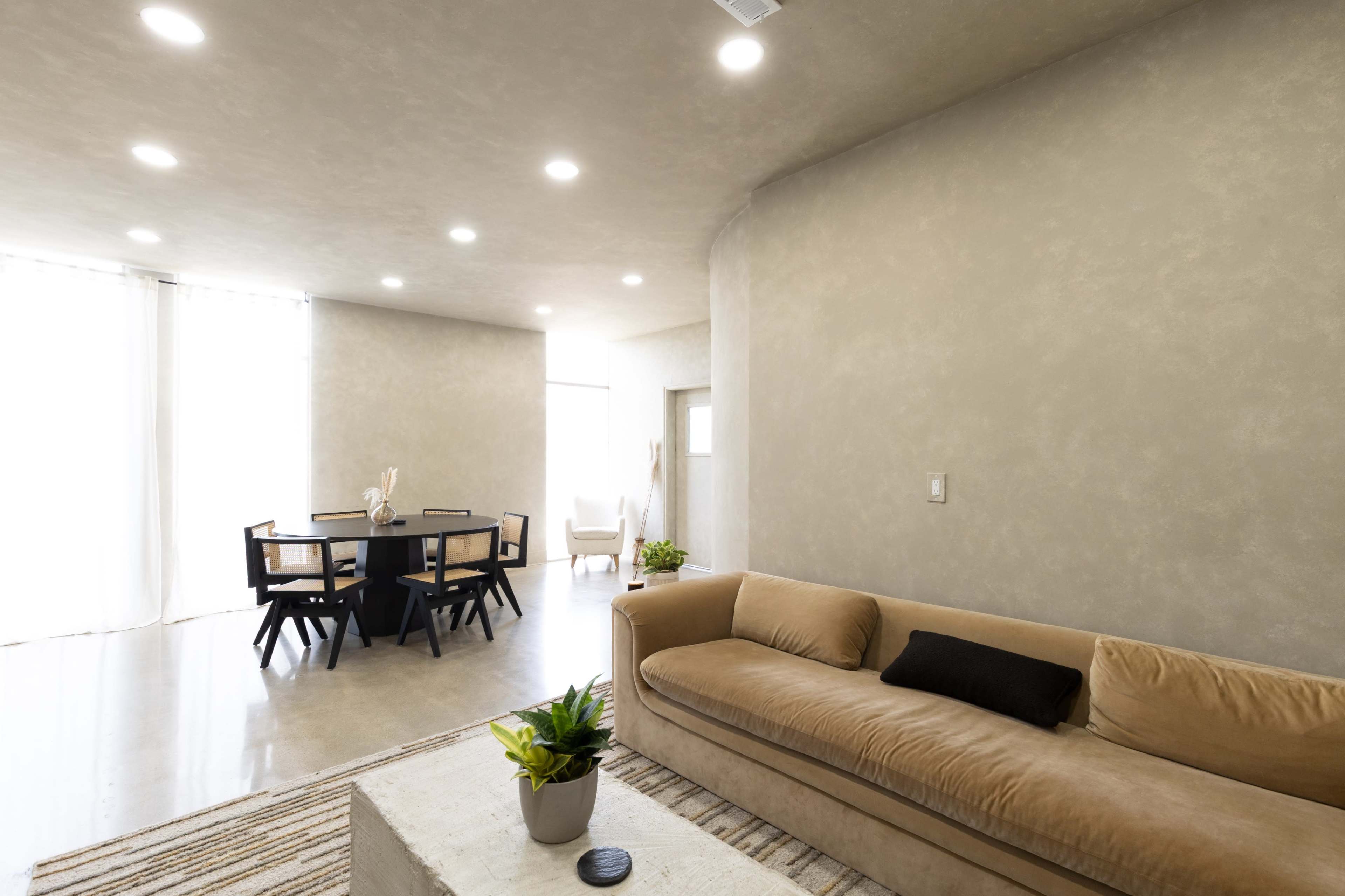 The image shows a modern living space featuring a beige sofa, a round dining table with black chairs, and large windows that allow natural light to enter the room.