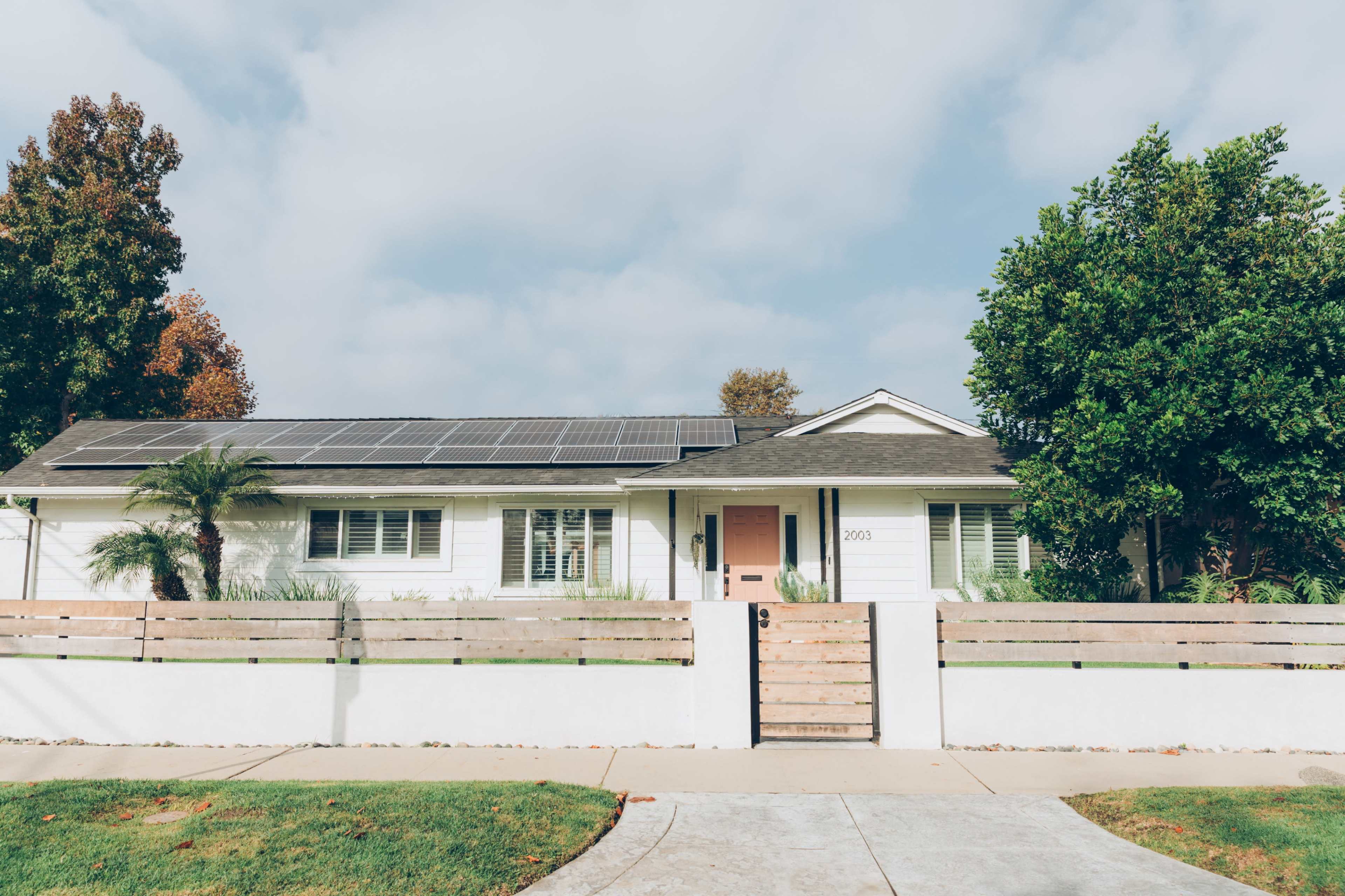 A single-story white house with solar panels on the roof is bordered by a wooden fence and a concrete pathway.