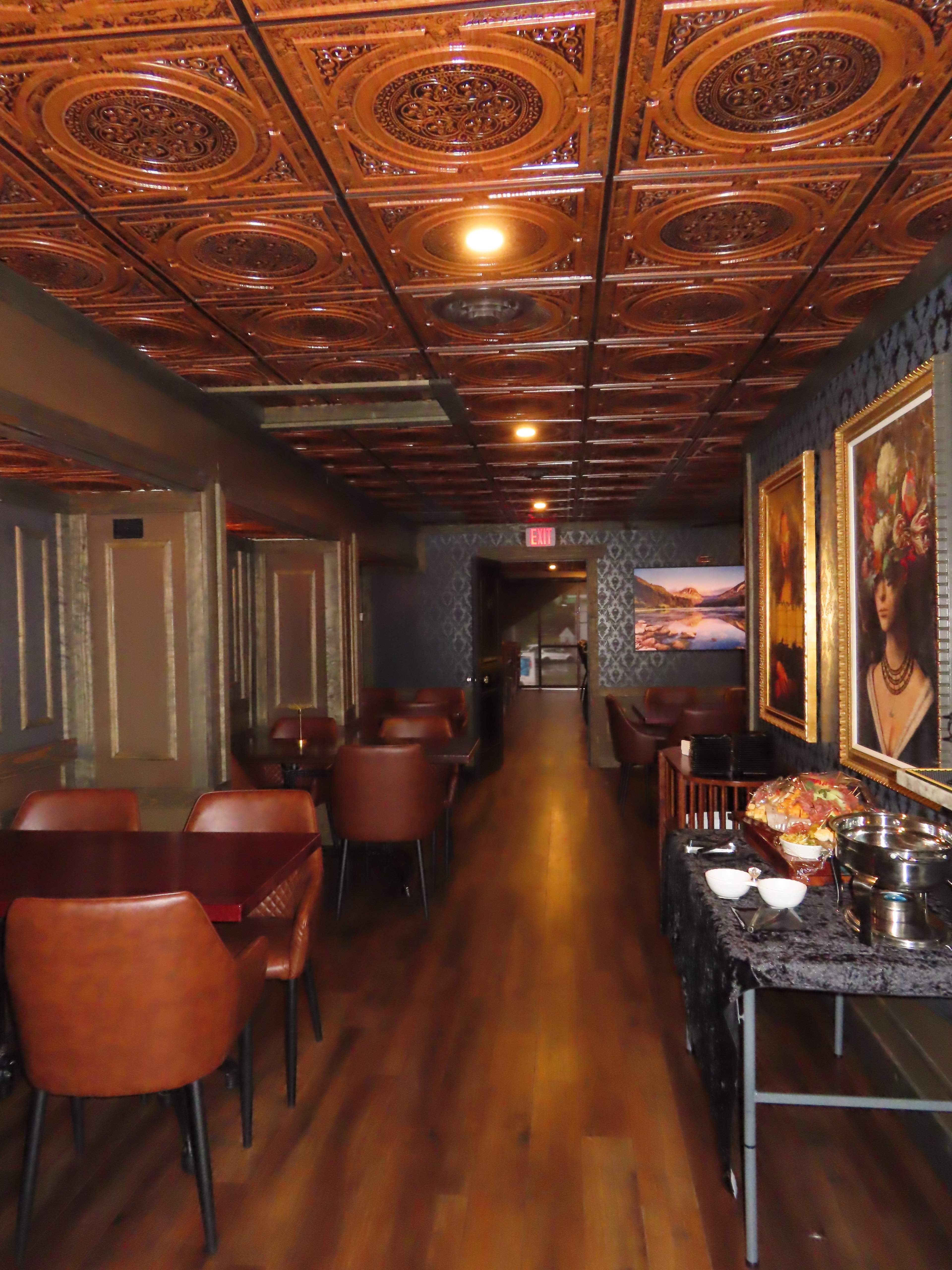 The image shows a dimly lit restaurant hallway with brown chairs, elaborately patterned wooden ceiling tiles, and decorative artwork on the walls.