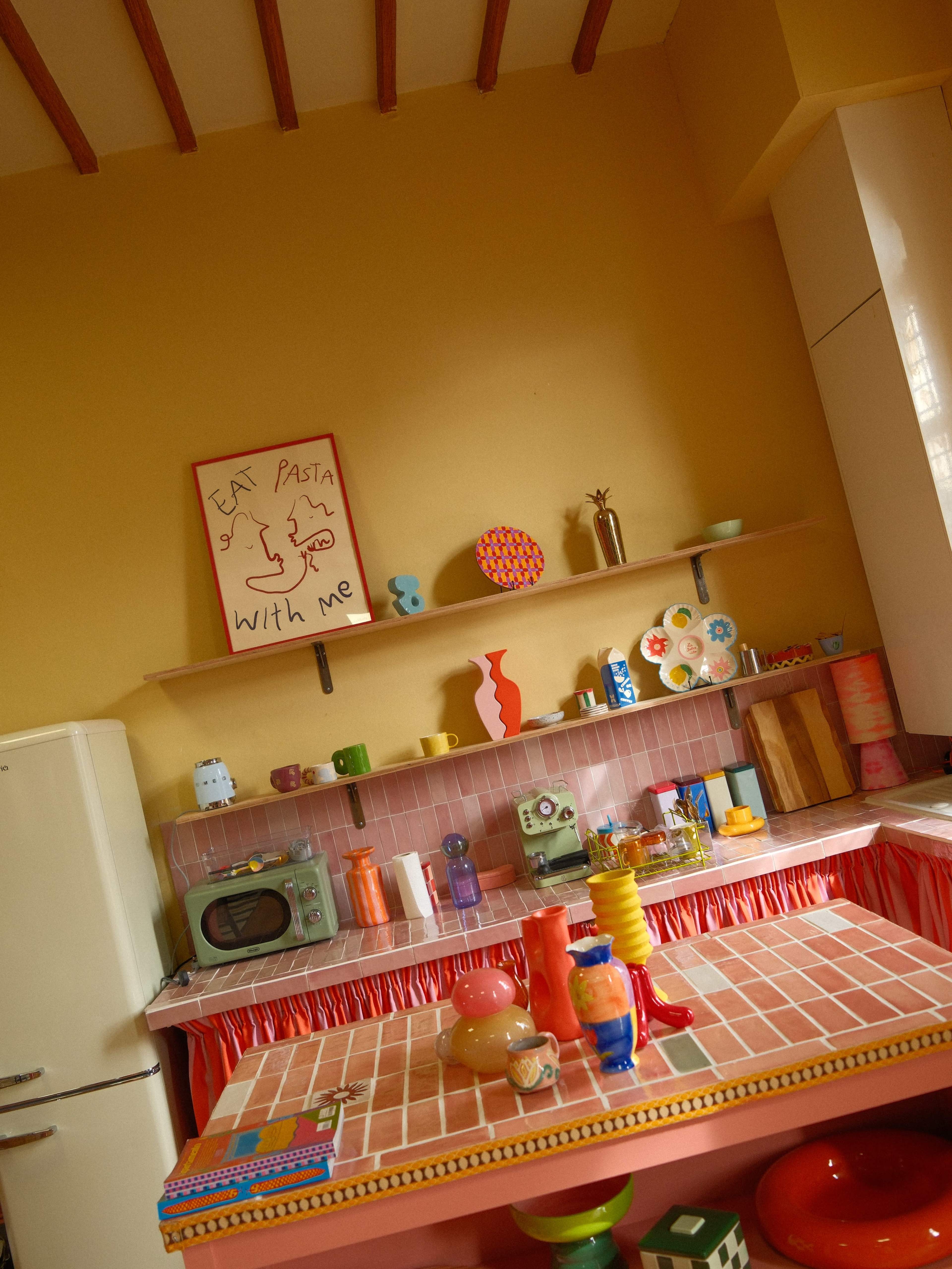 The image shows a brightly colored kitchen with a tiled countertop, retro appliances, and decorative items arranged on shelves.