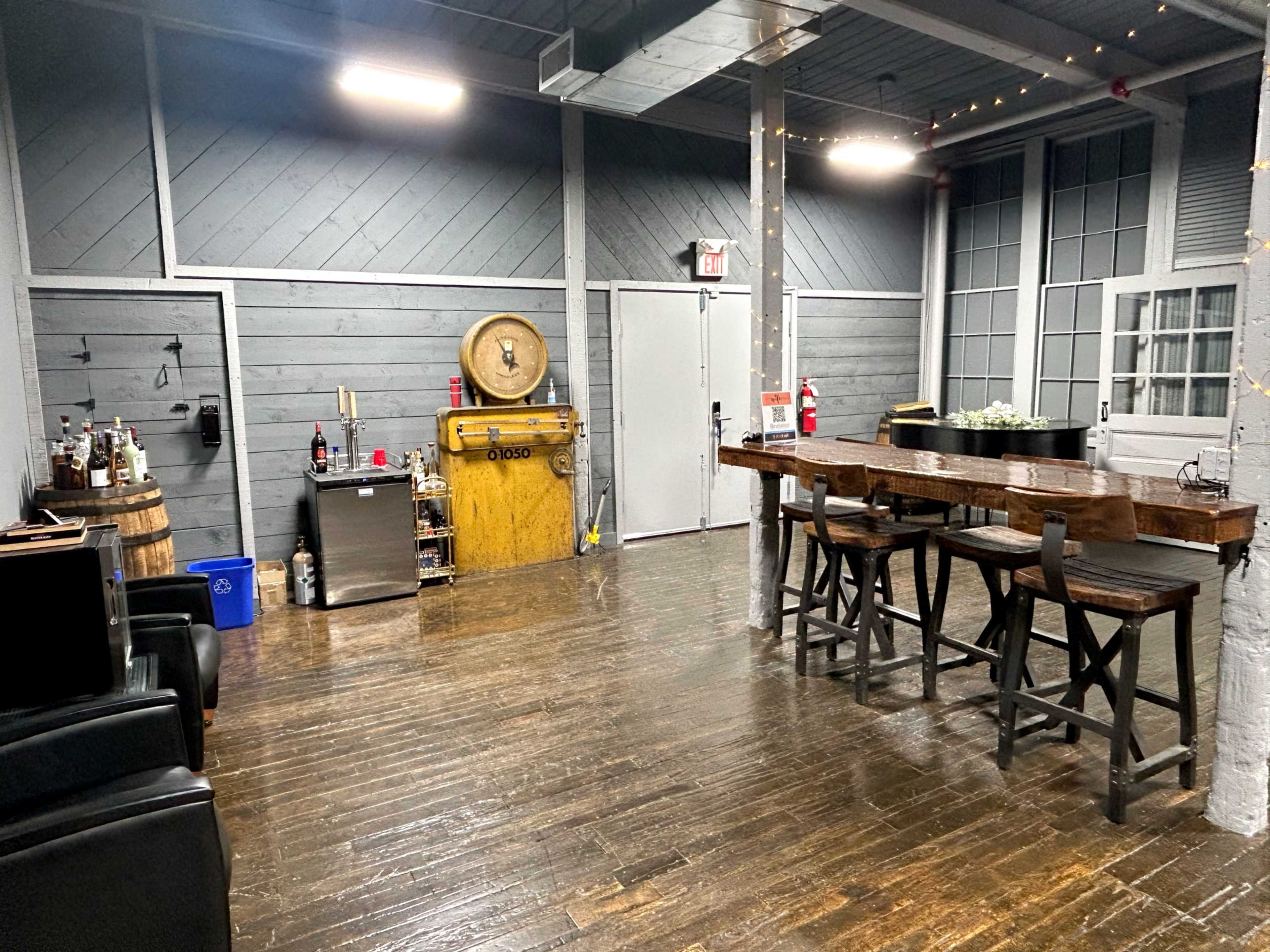 The image shows an interior space with dark wooden flooring, a bar with high stools, a fridge, and various decorations, including a barrel and a yellow cabinet.