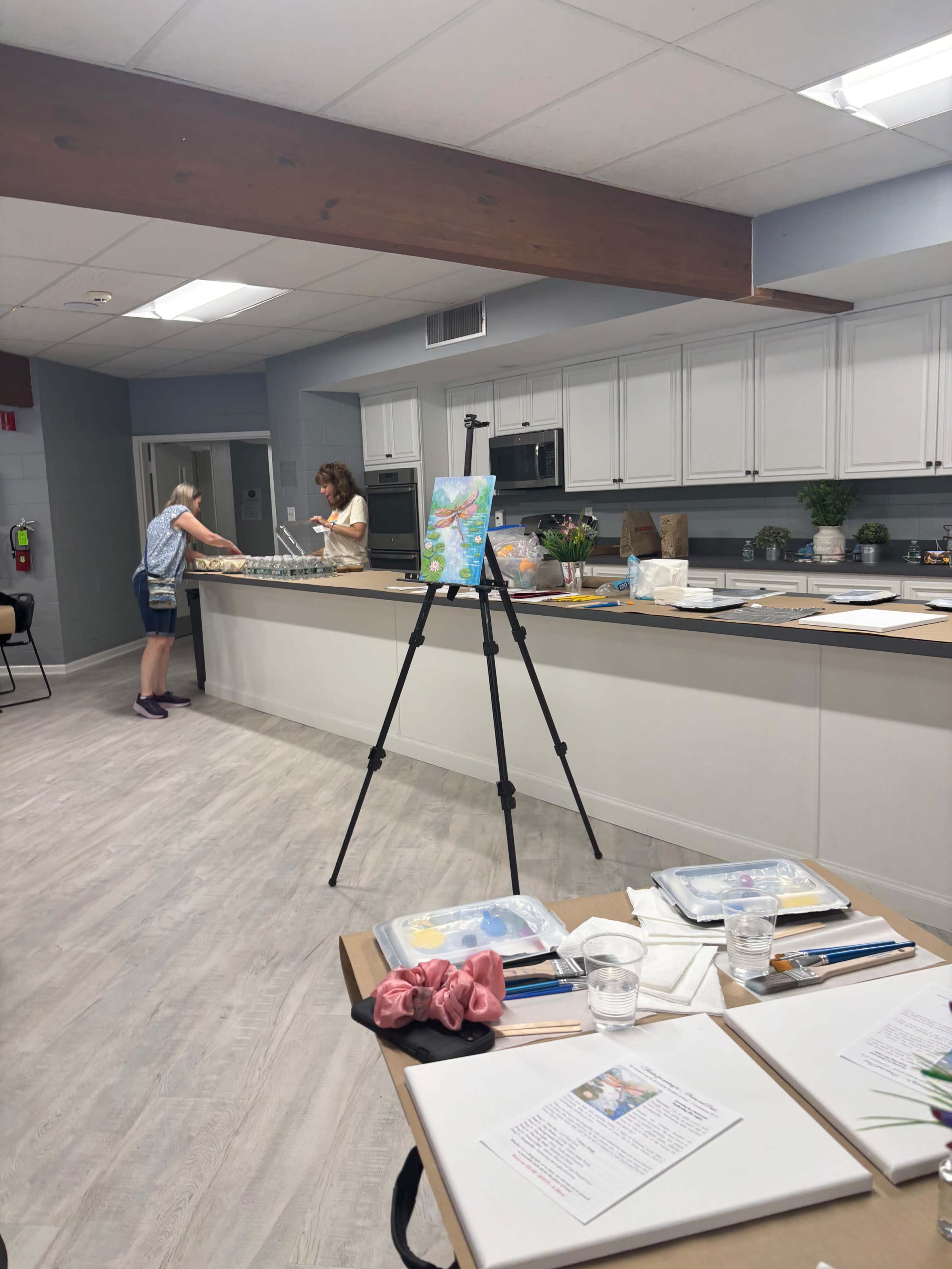 A spacious kitchen with a large island and an easel holding an artwork, where one person is working at the counter while another is in the background near the refrigerator.
