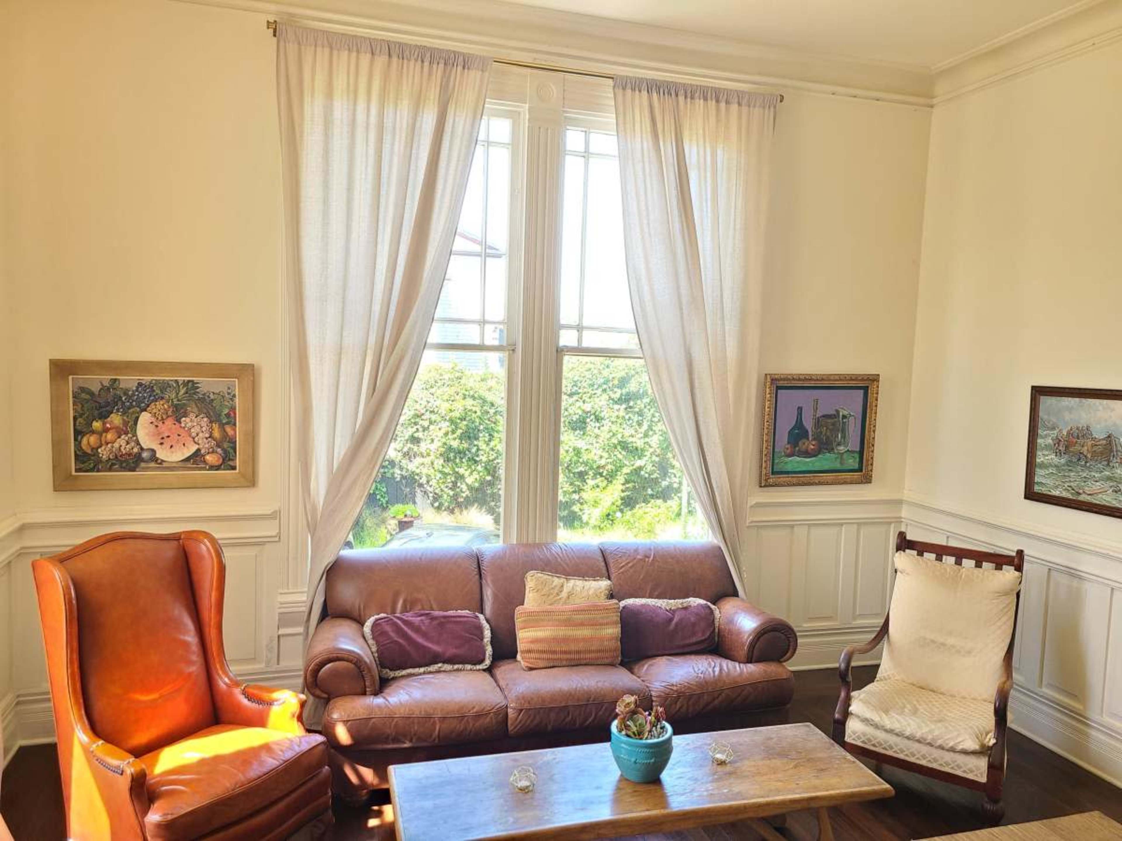 The room features a leather sofa, an armchair, and a rocking chair, with large windows letting in natural light and artwork hanging on the walls.