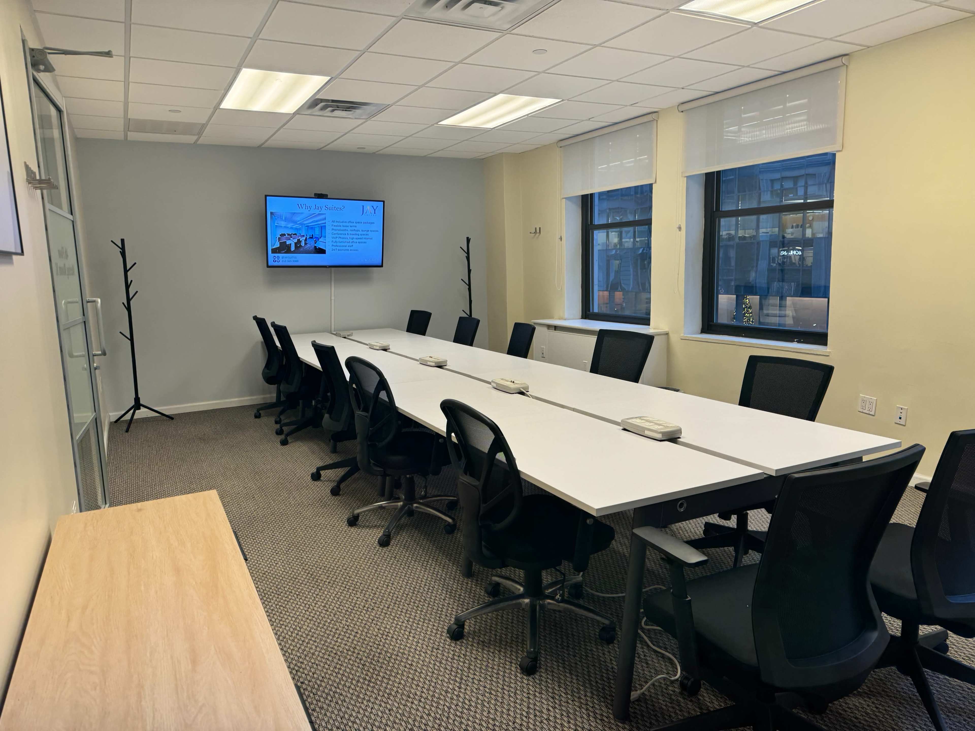 A conference room features a long table surrounded by black chairs, with a television screen displaying information on the wall.