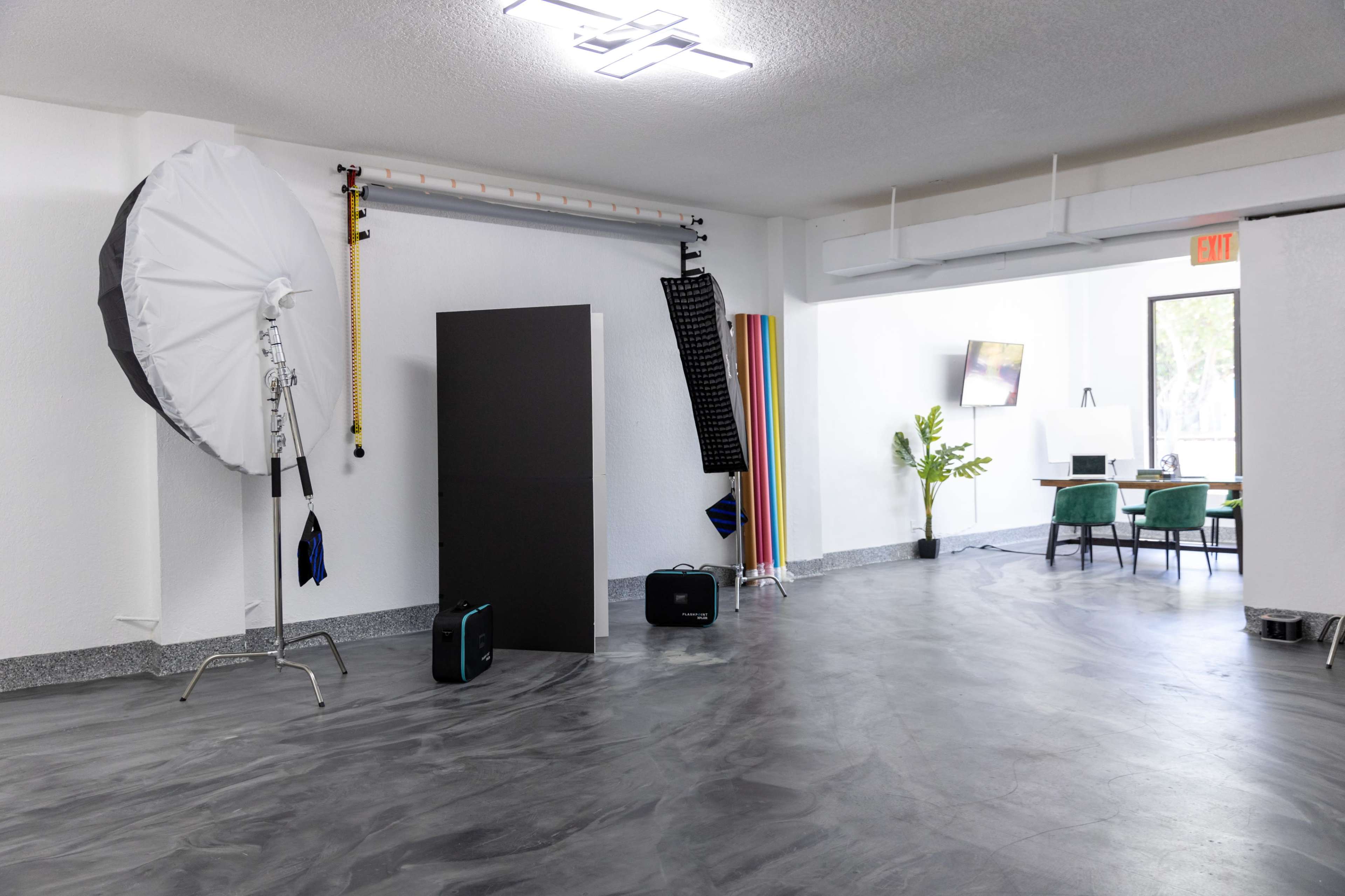 A well-lit studio space with photography equipment, including an umbrella softbox, a black backdrop, and a variety of colored boards, alongside a workspace with a table and chairs.
