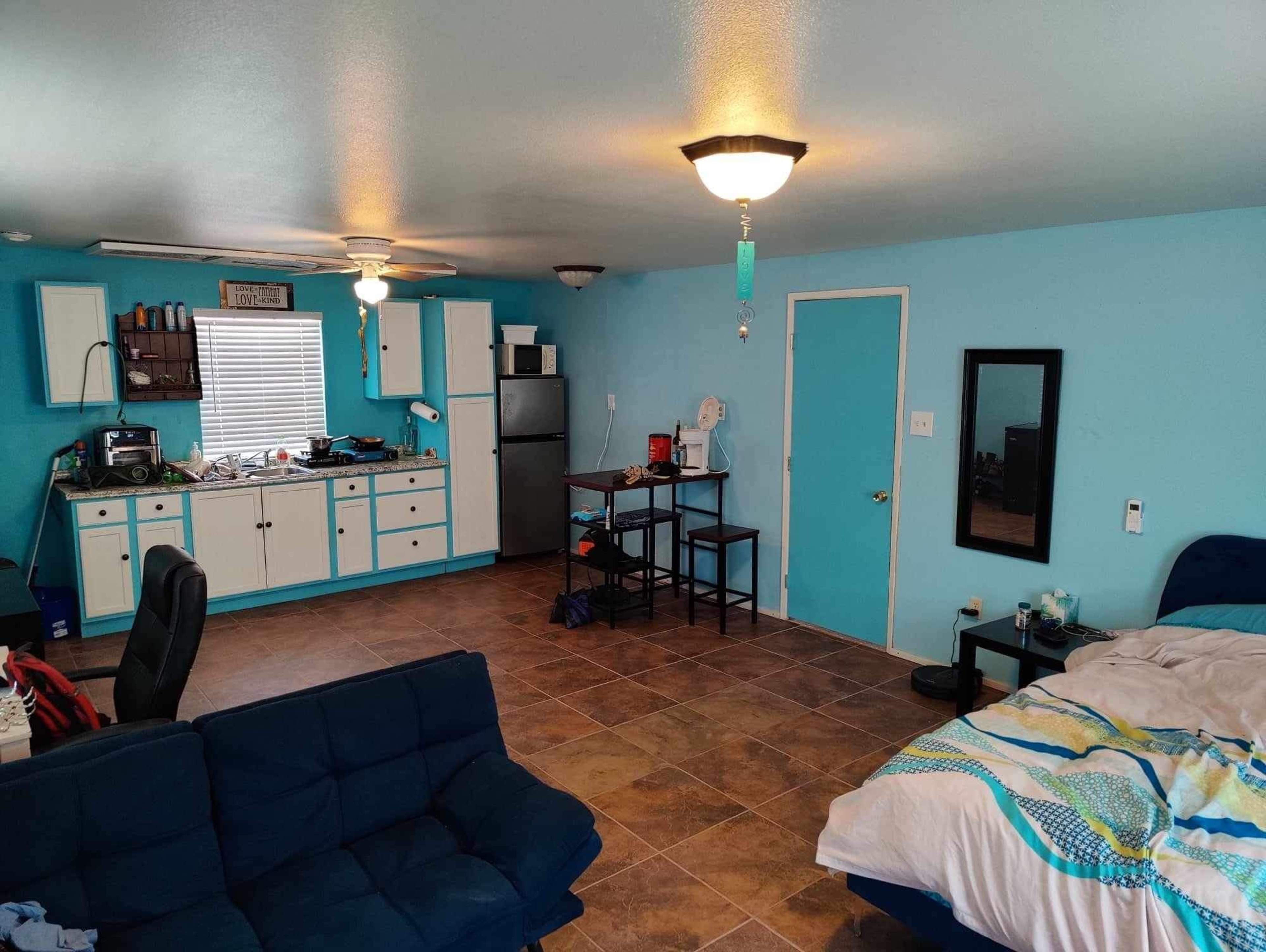 The image shows a small, open living space with a kitchen area, a dining table, a couch, and a bed, all featuring light blue walls and tiled flooring.