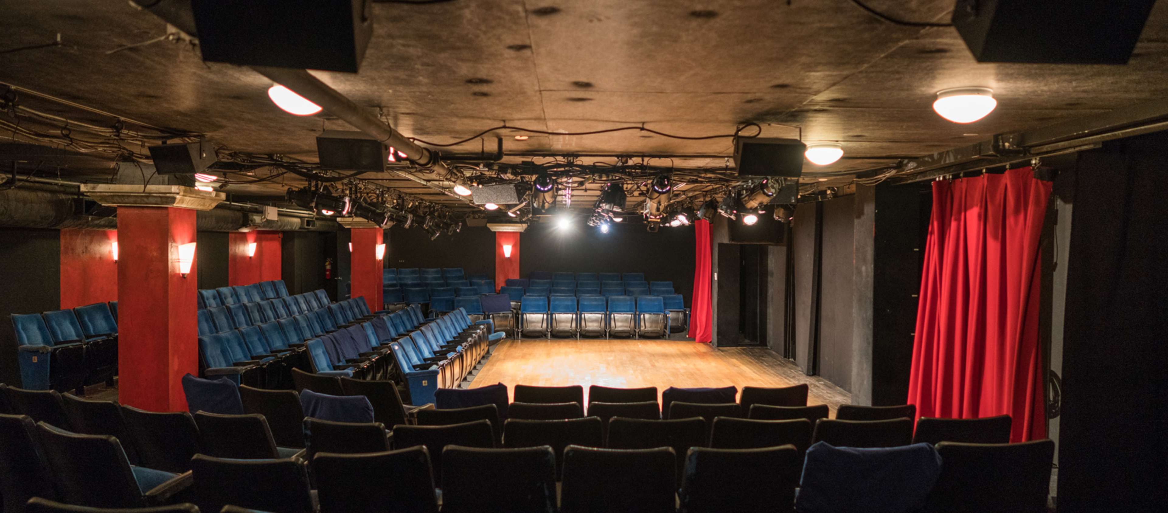 A small theater with blue seating arranged in rows facing a wooden stage, flanked by red curtains and overhead lights.