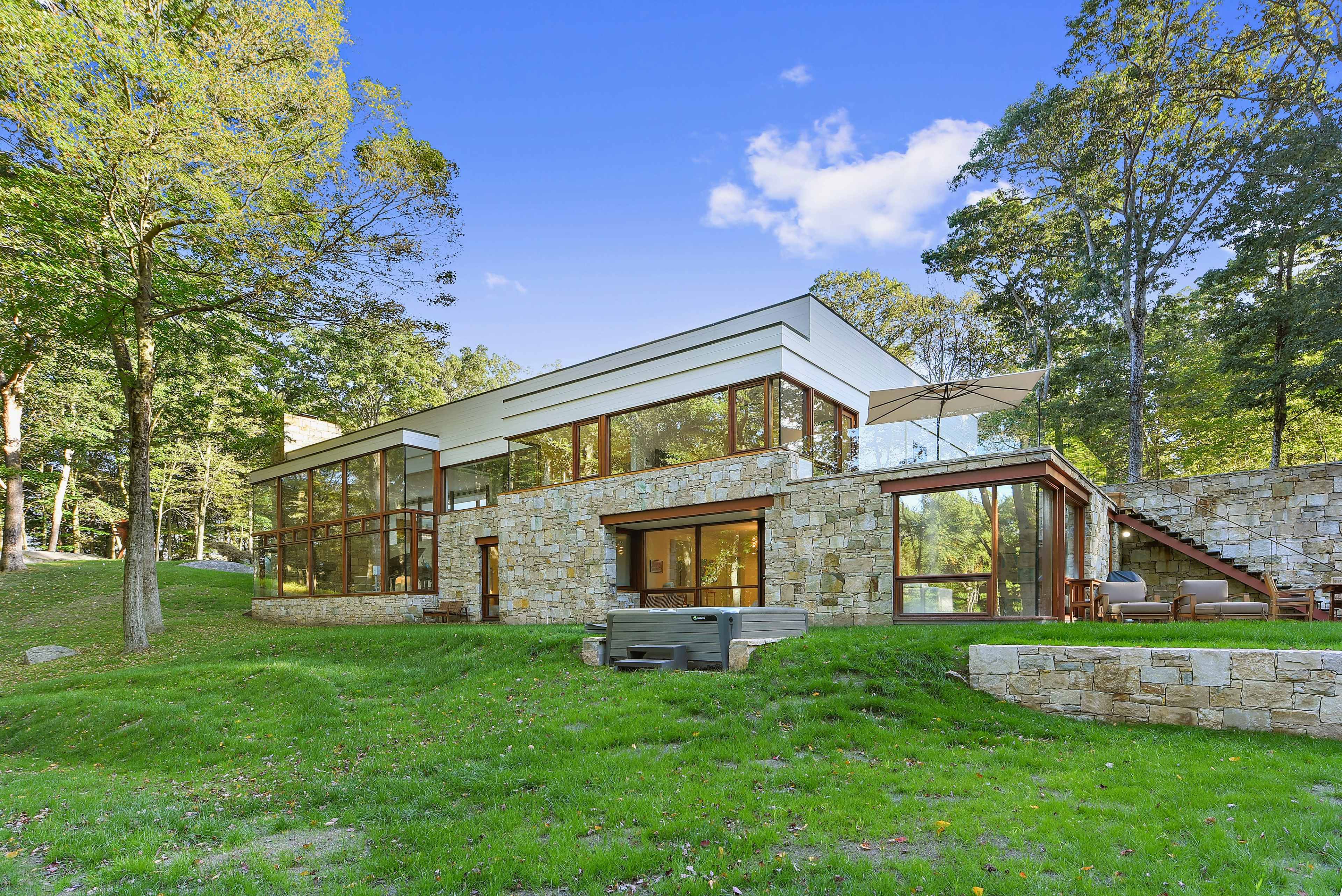 The image shows a modern house with large glass windows and stone accents, situated on a grassy landscape surrounded by trees.