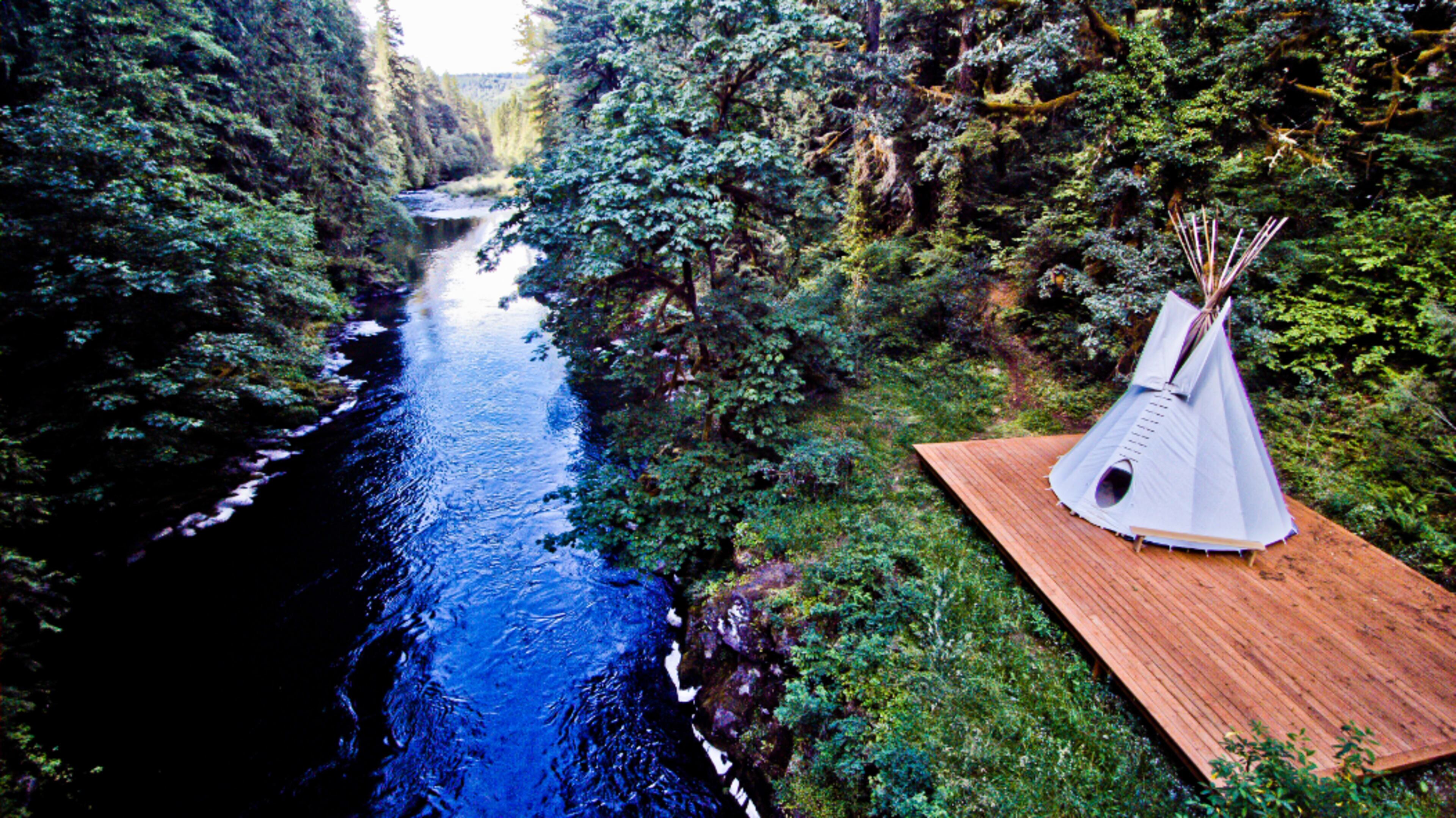 A traditional teepee is set on a wooden platform beside a flowing river, surrounded by dense greenery and tall trees.