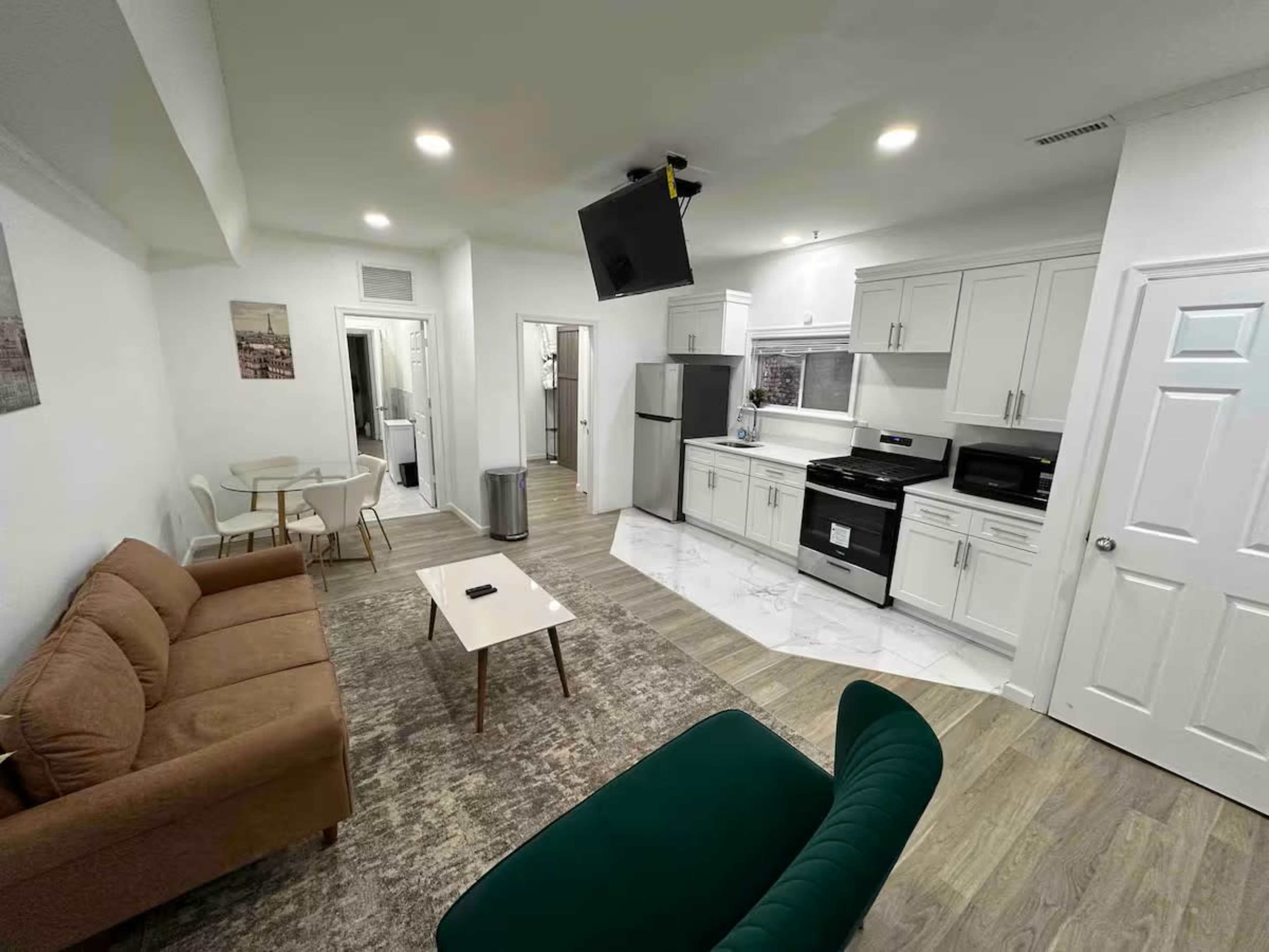 The image shows a modern living space featuring a kitchen with white cabinets and appliances, a brown couch, a green chair, and a dining table.