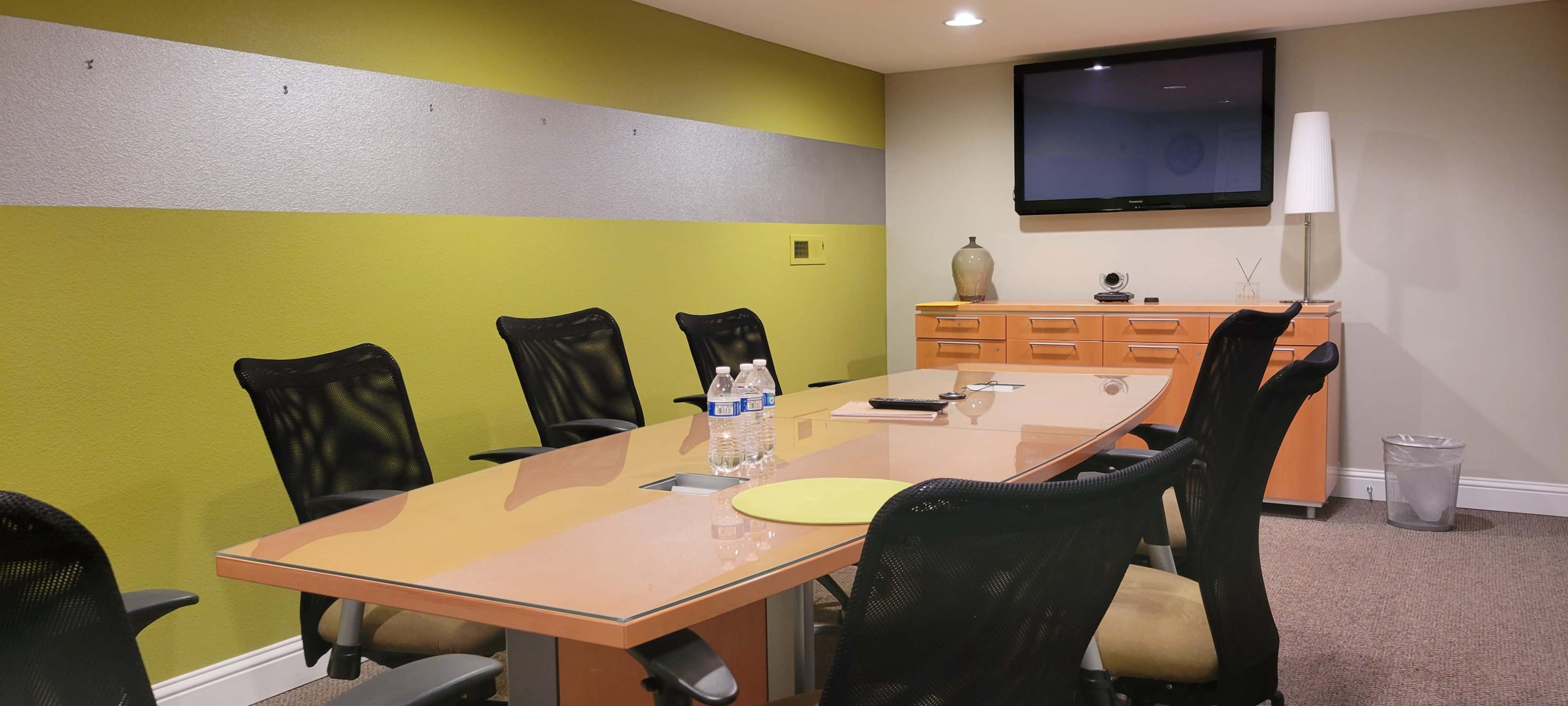 A modern conference room features a large table surrounded by black chairs, with a television mounted on the wall and a side cabinet.