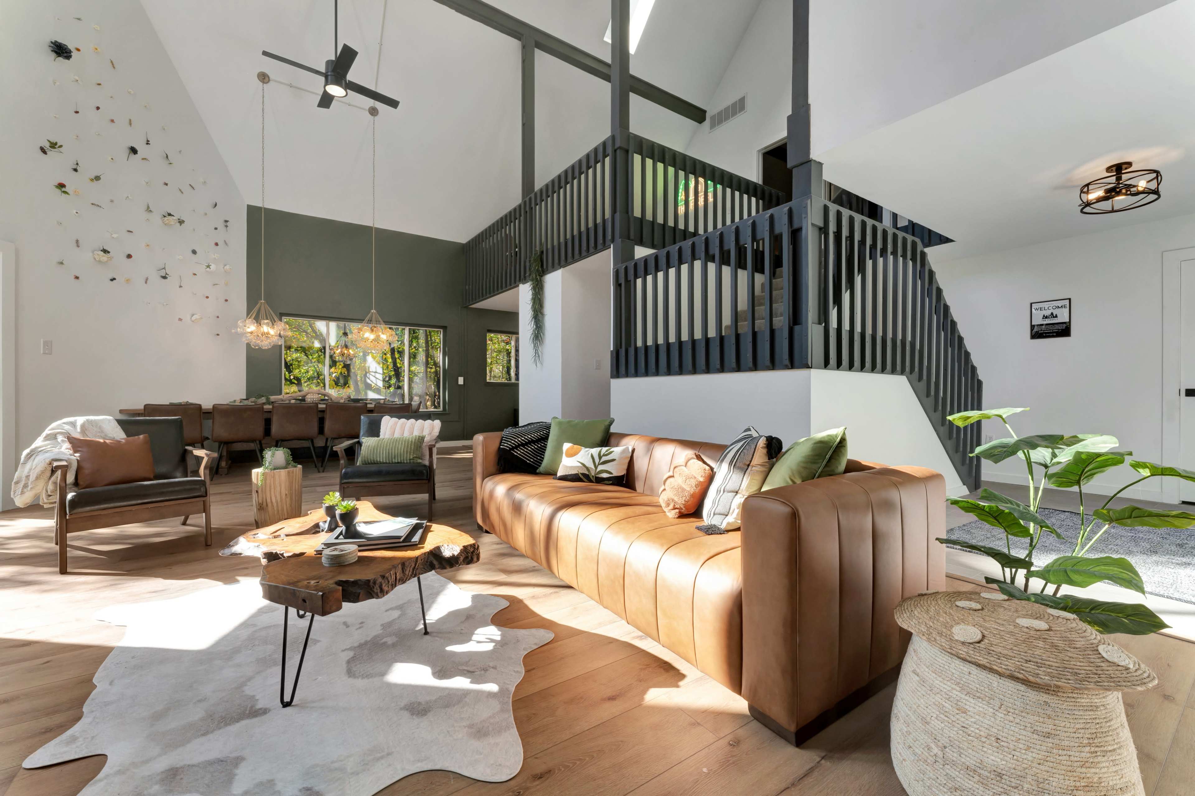 The image shows a modern living room with a brown leather sofa, a wooden coffee table, and a staircase leading to an upper area, surrounded by large windows.