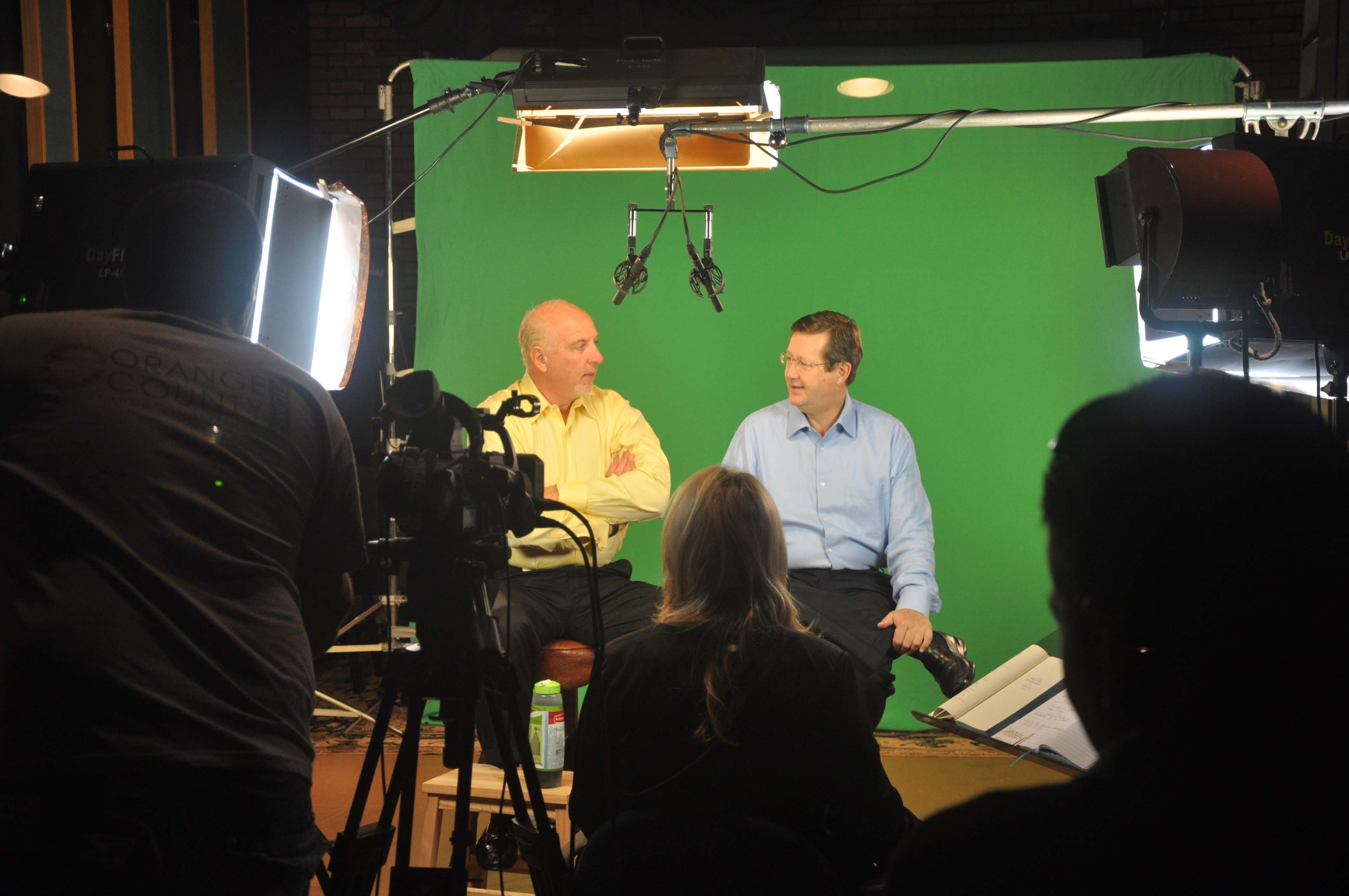 Two men sit on chairs in front of a green screen, while a crew prepares filming equipment in a studio setting.