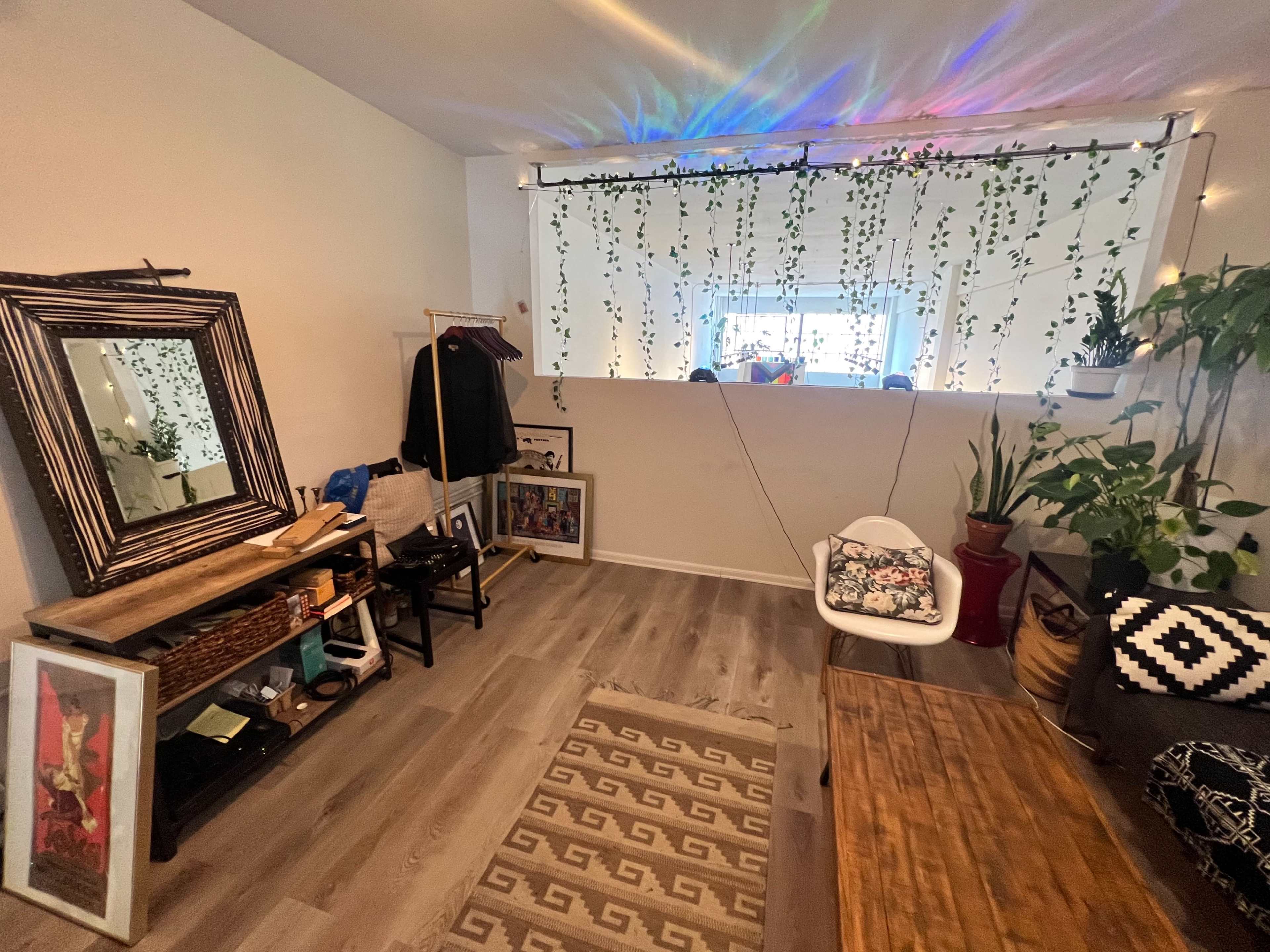 A cozy, well-decorated room with a large mirror, various plants, artwork on the walls, and a wooden table.