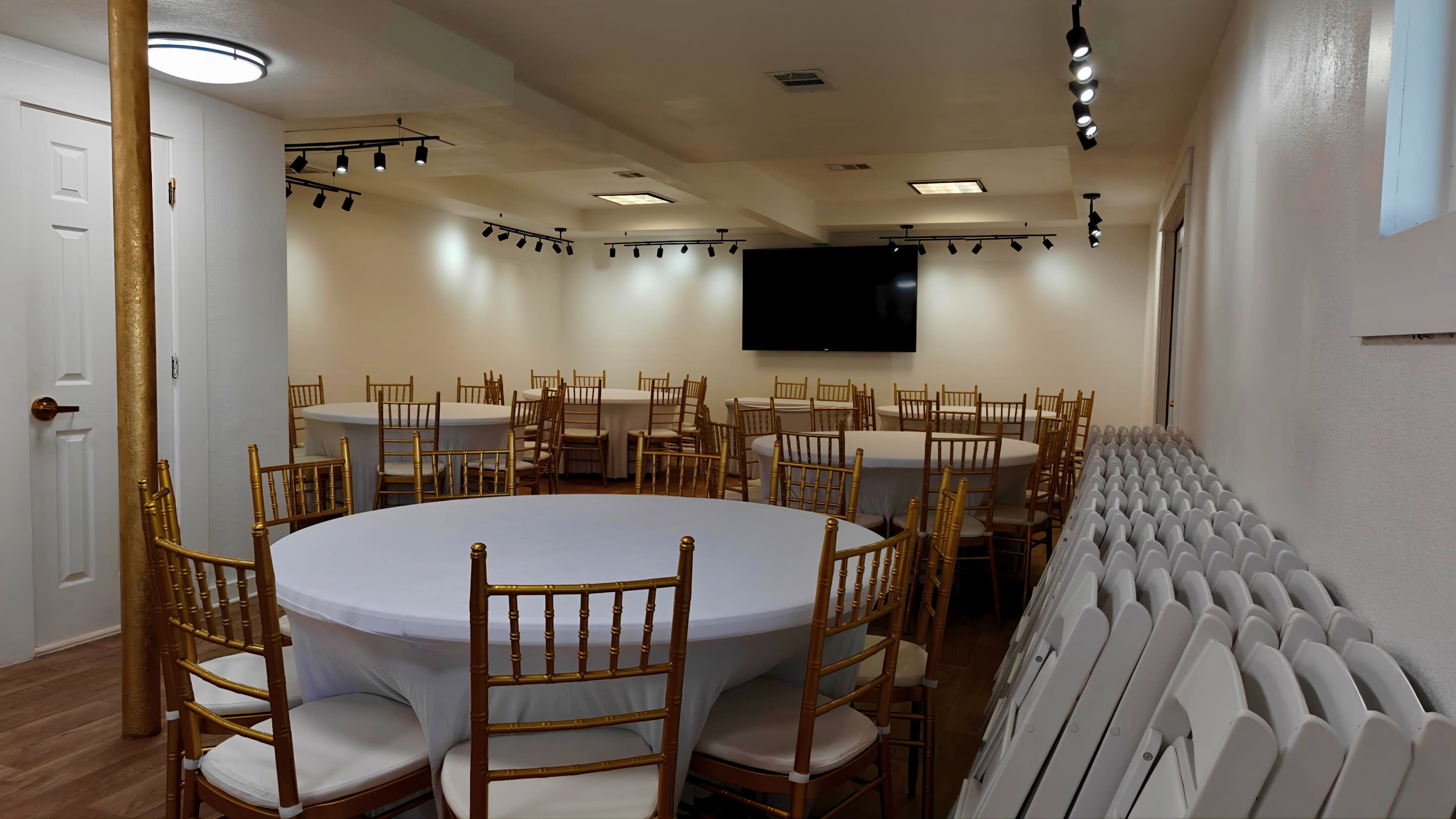 The image shows a well-lit event space with several round tables covered in white tablecloths and gold chiavari chairs arranged throughout the room.