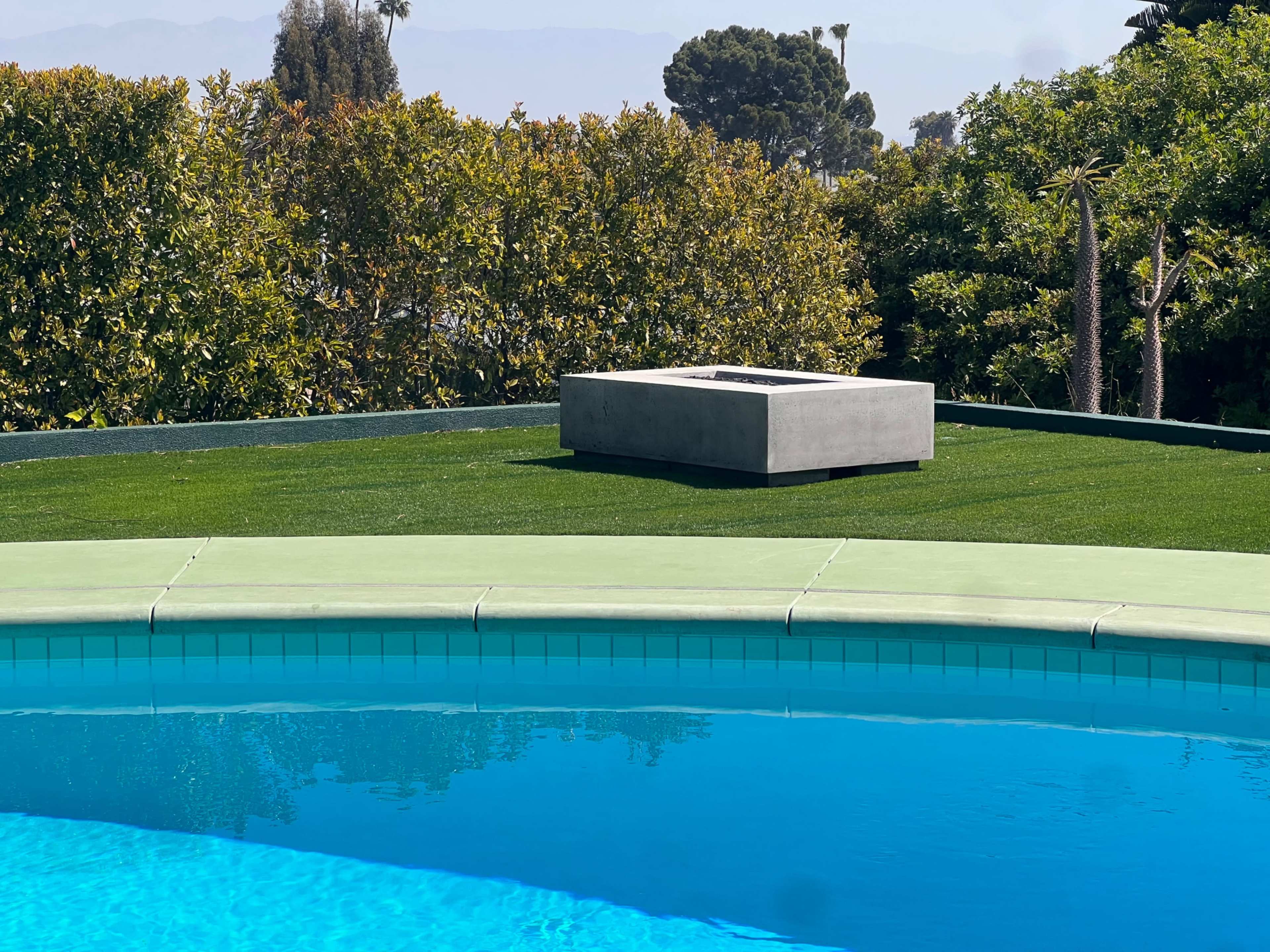 A modern concrete fire pit is situated on a grassy area overlooking a blue swimming pool.