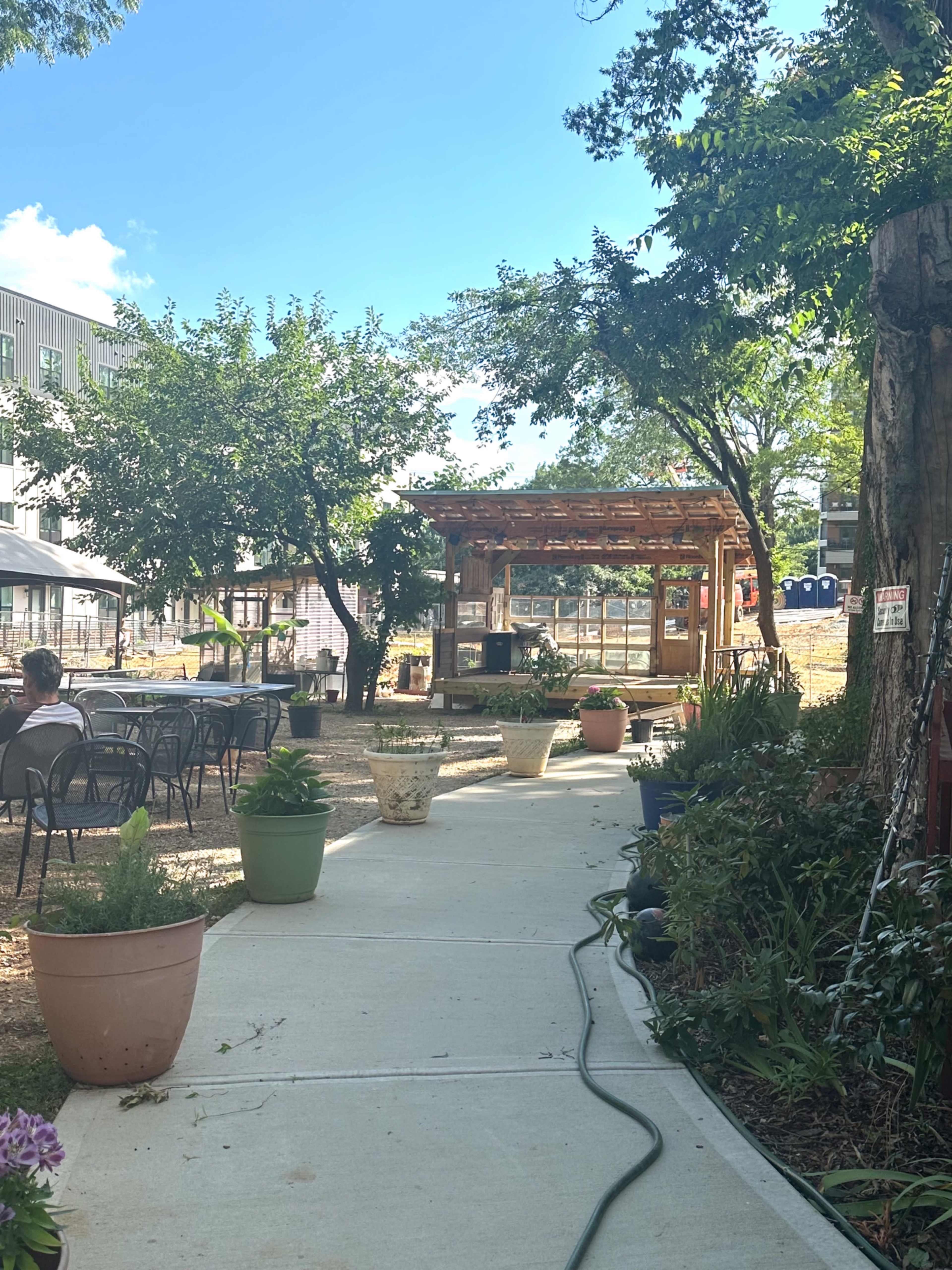A paved walkway leads through a garden area with various potted plants and seating, ending at a wooden pavilion surrounded by trees.