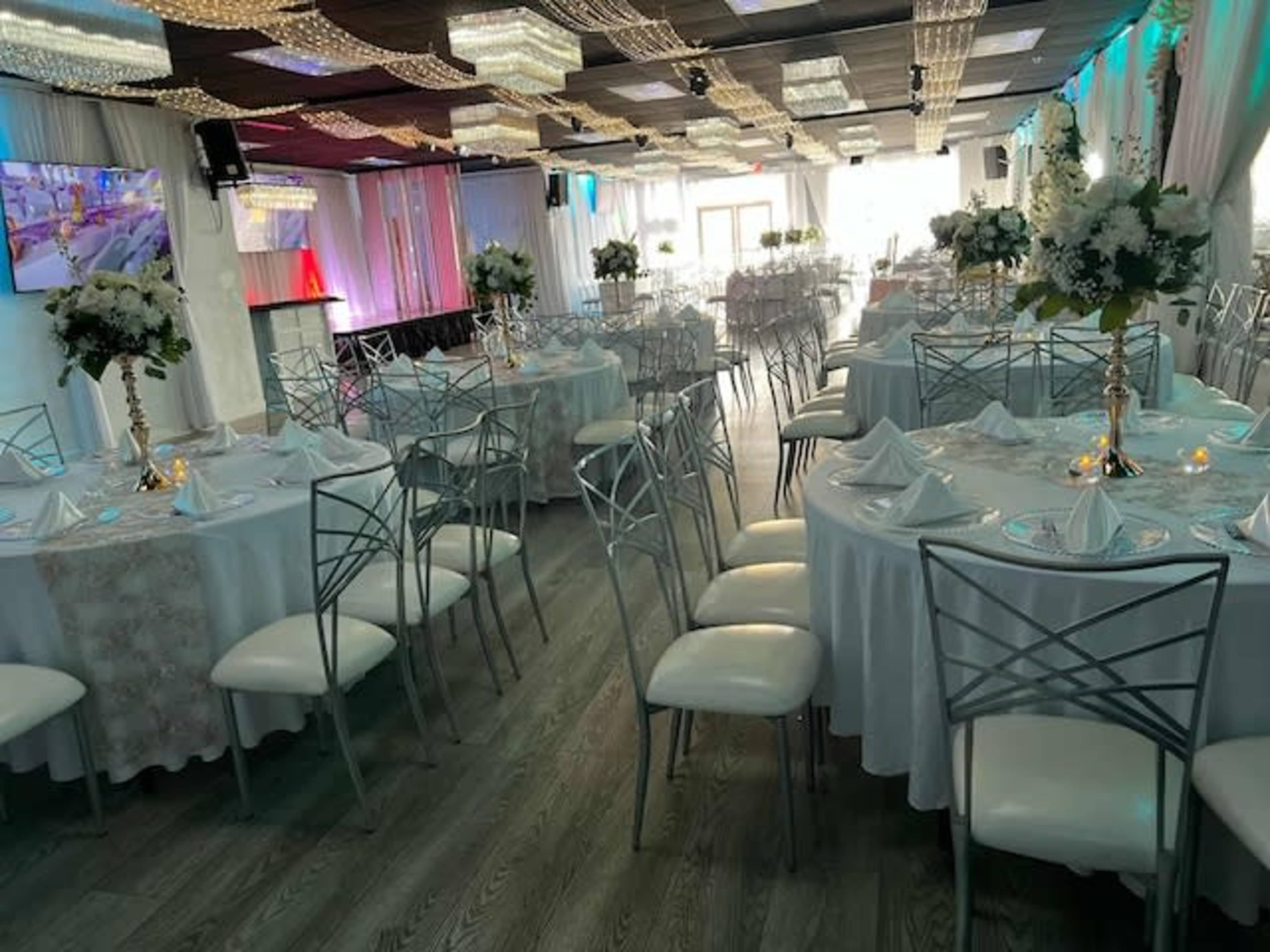 The image shows an elegantly set banquet hall with round tables covered in white tablecloths, each adorned with floral centerpieces and neatly arranged napkins.