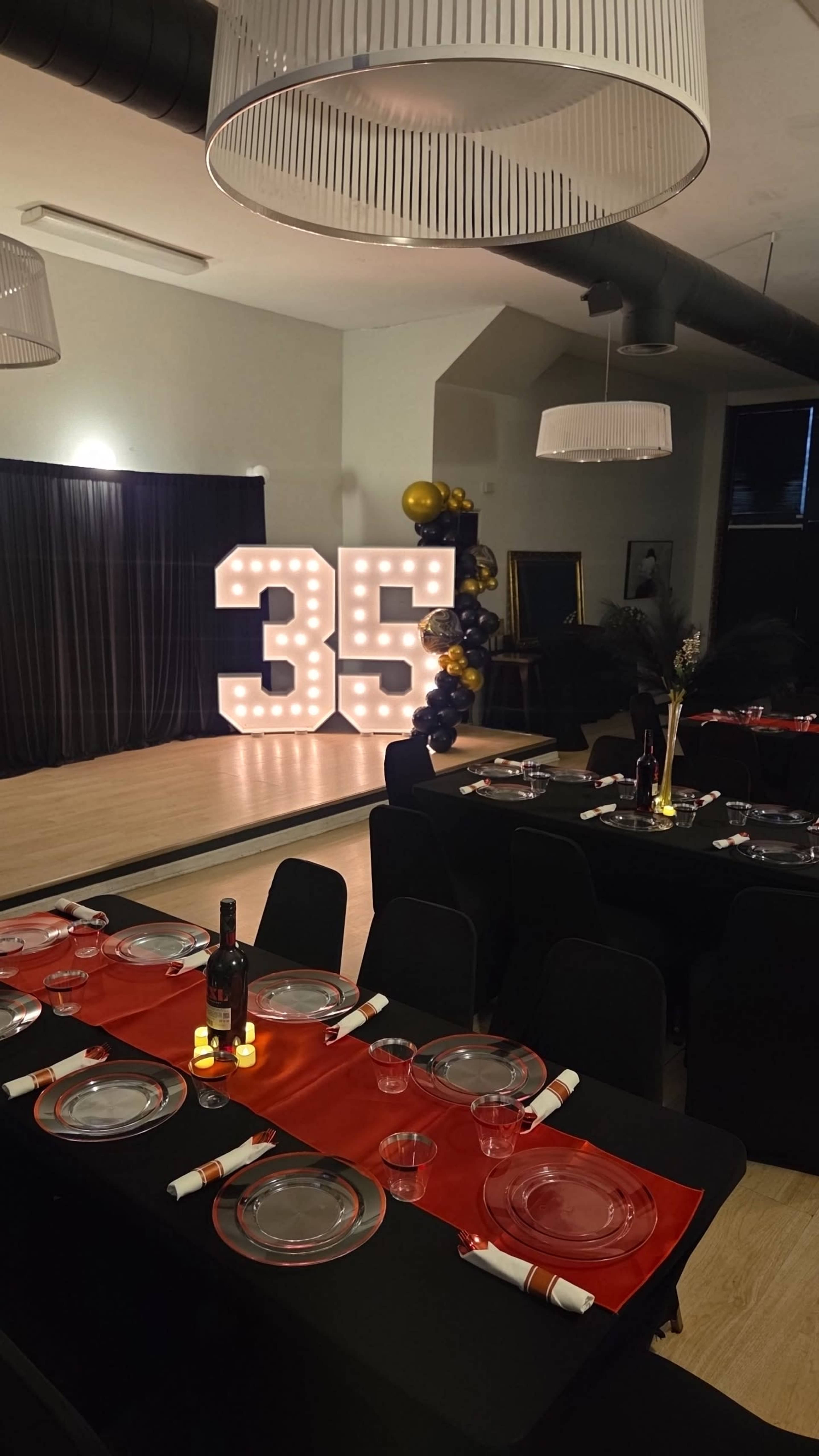A party setup features a large illuminated "35" sign against a backdrop of black curtains, with tables set for guests, decorated with red and black tablecloths.