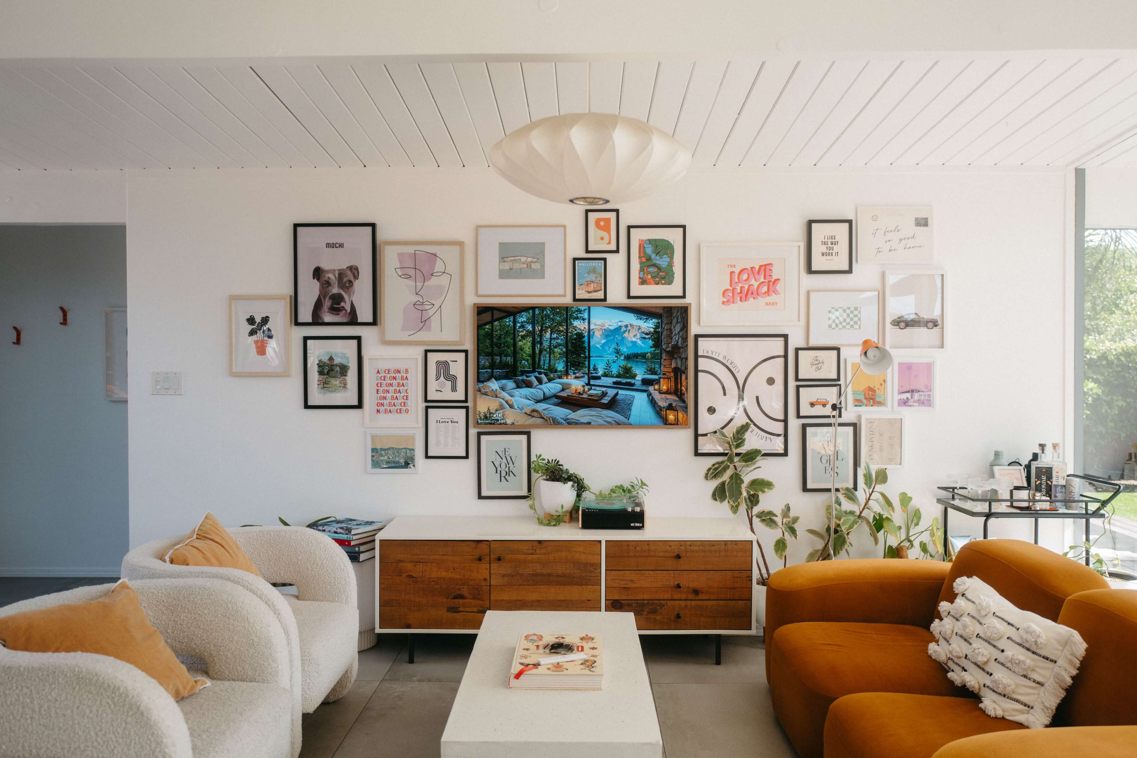 A cozy living room features a wall adorned with various framed artworks, a television mounted on a light-colored wall, and stylish furniture including a white and orange sofa set.
