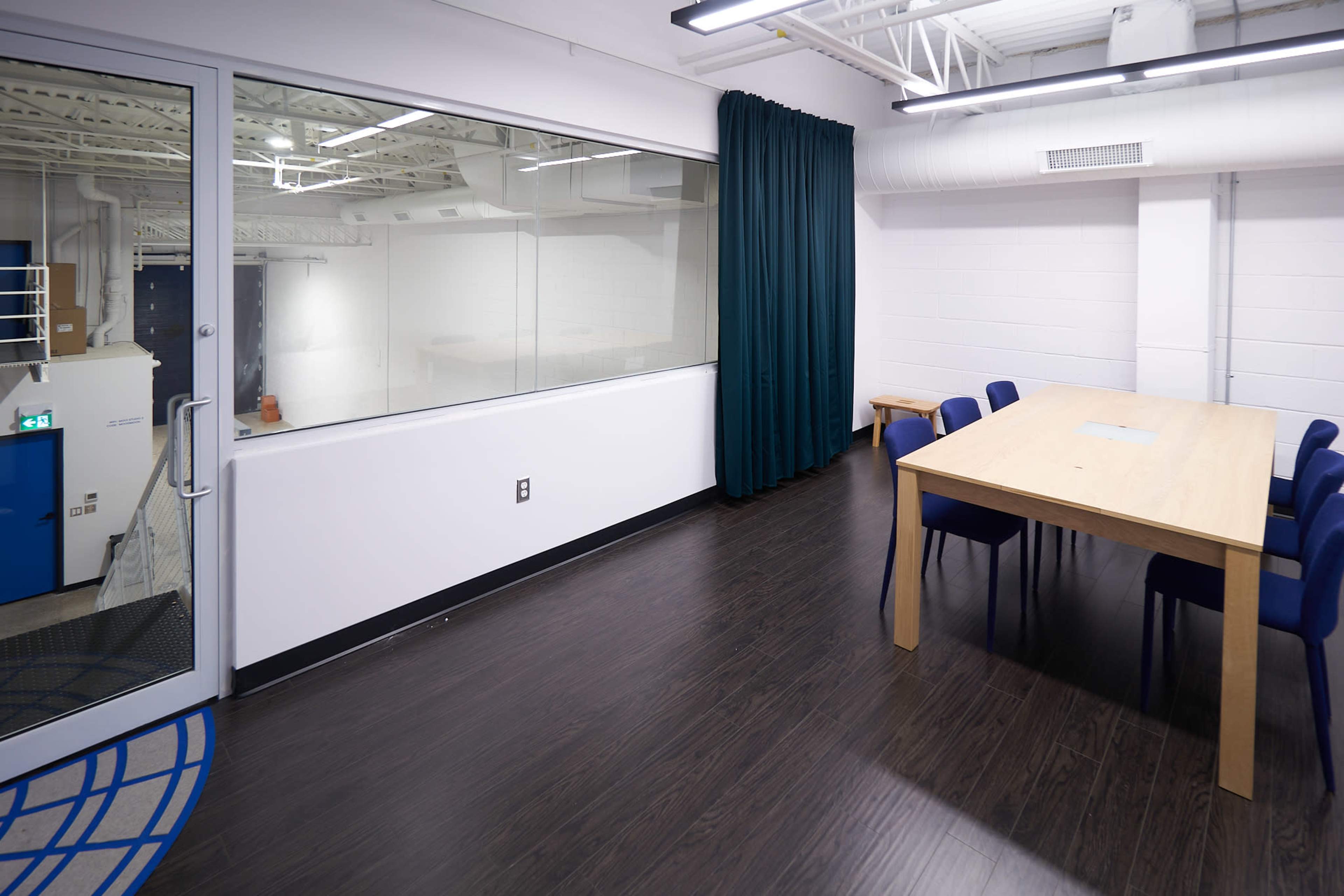 A modern meeting room with a large wooden table and blue chairs, featuring a glass wall that overlooks a separate space.