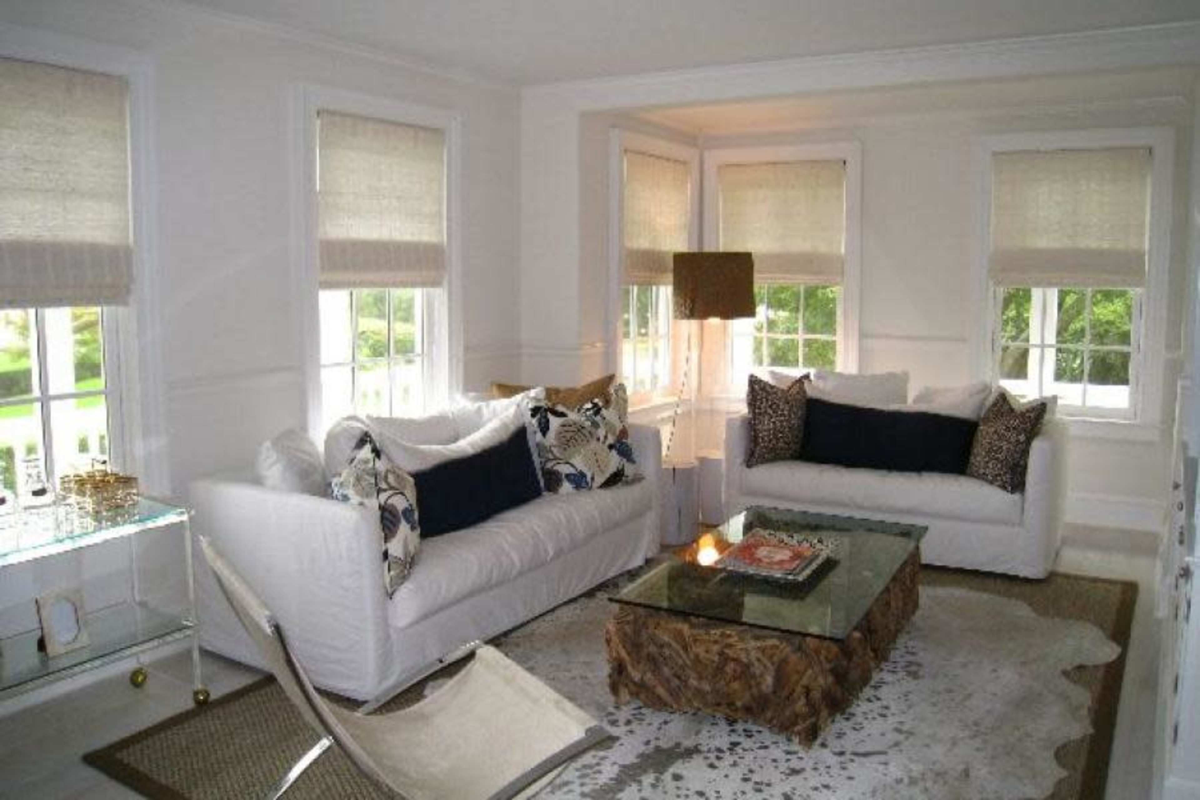 A light-filled living room features two white sofas flanking a glass coffee table on a patterned rug, with large windows and neutral window shades.
