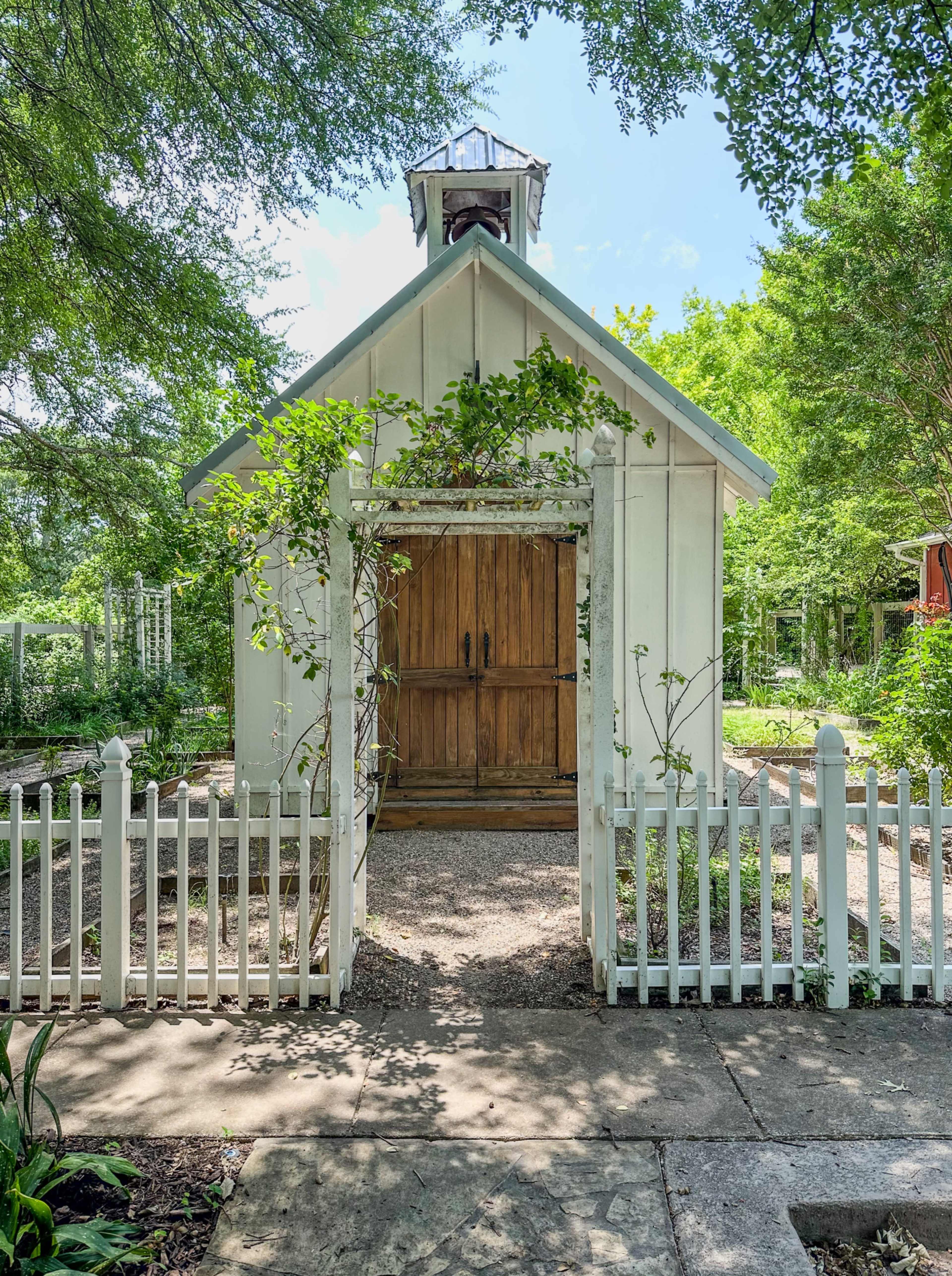 A small, white garden shed with a wooden door and a bell tower is surrounded by a picket fence and greenery.
