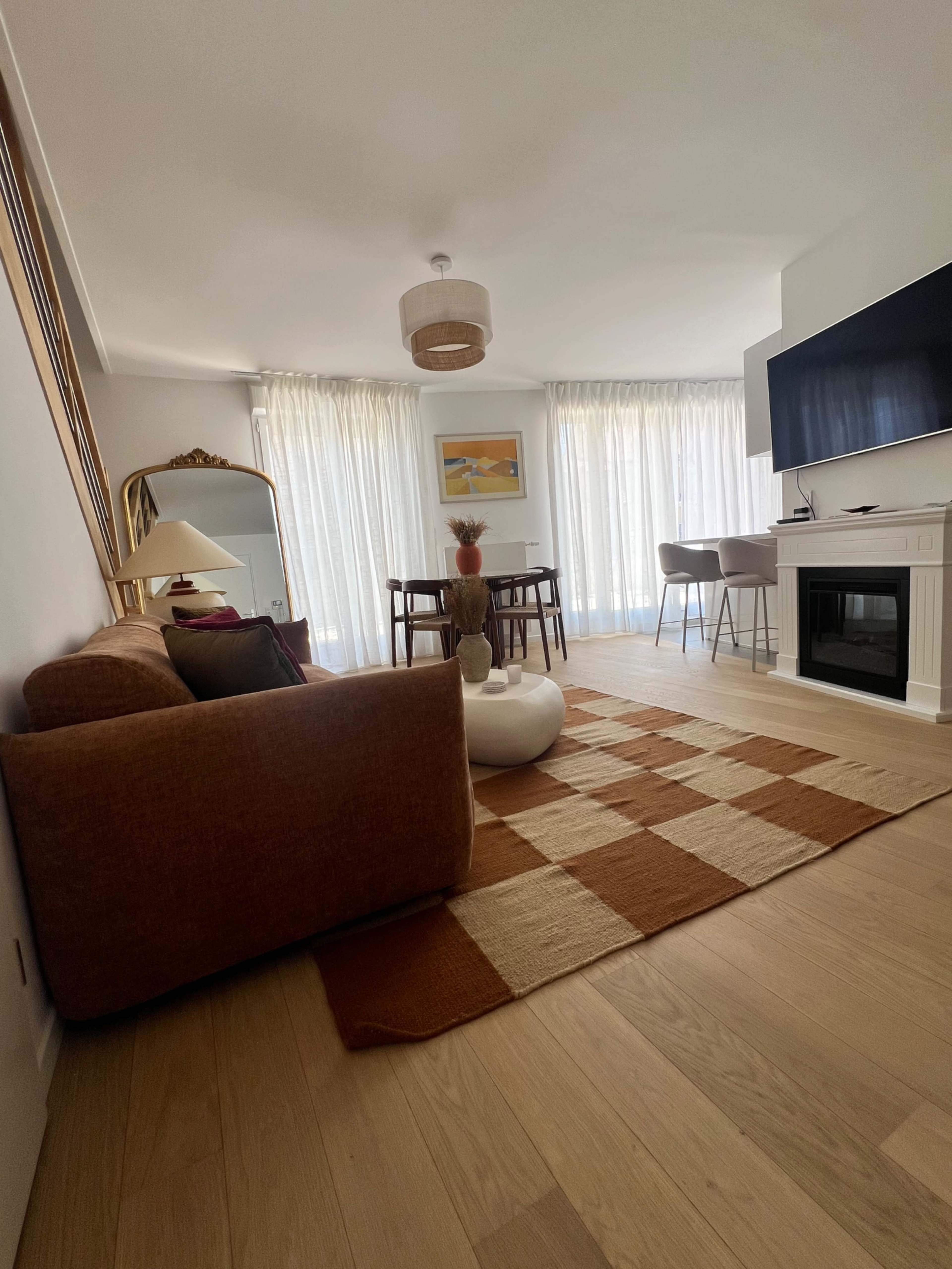 The image portrays a modern living room with a brown sofa, a round coffee table, and large windows with sheer curtains.