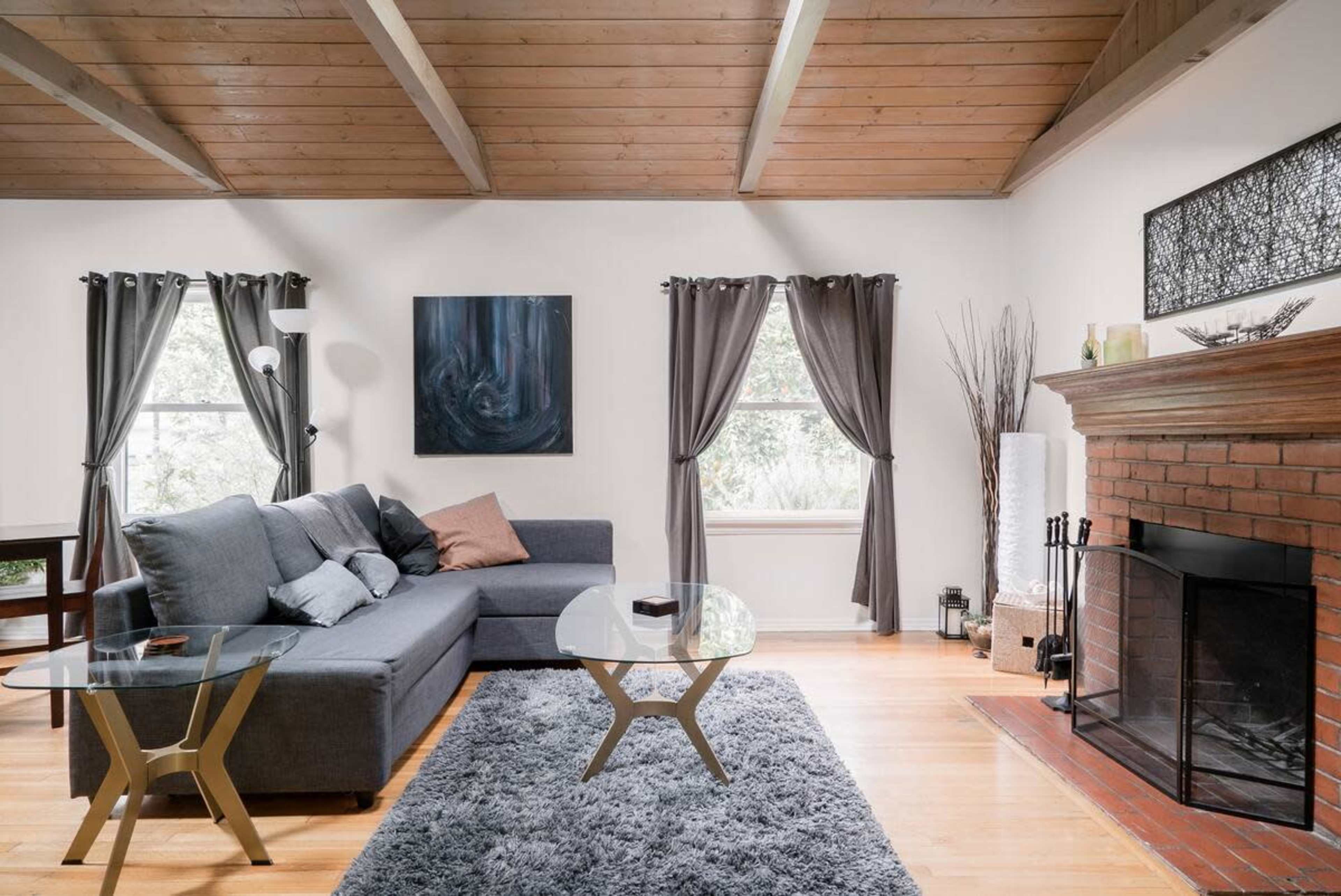 The living room features a gray sectional sofa, a glass coffee table, and a brick fireplace, all under a wooden ceiling with large windows dressed in dark curtains.
