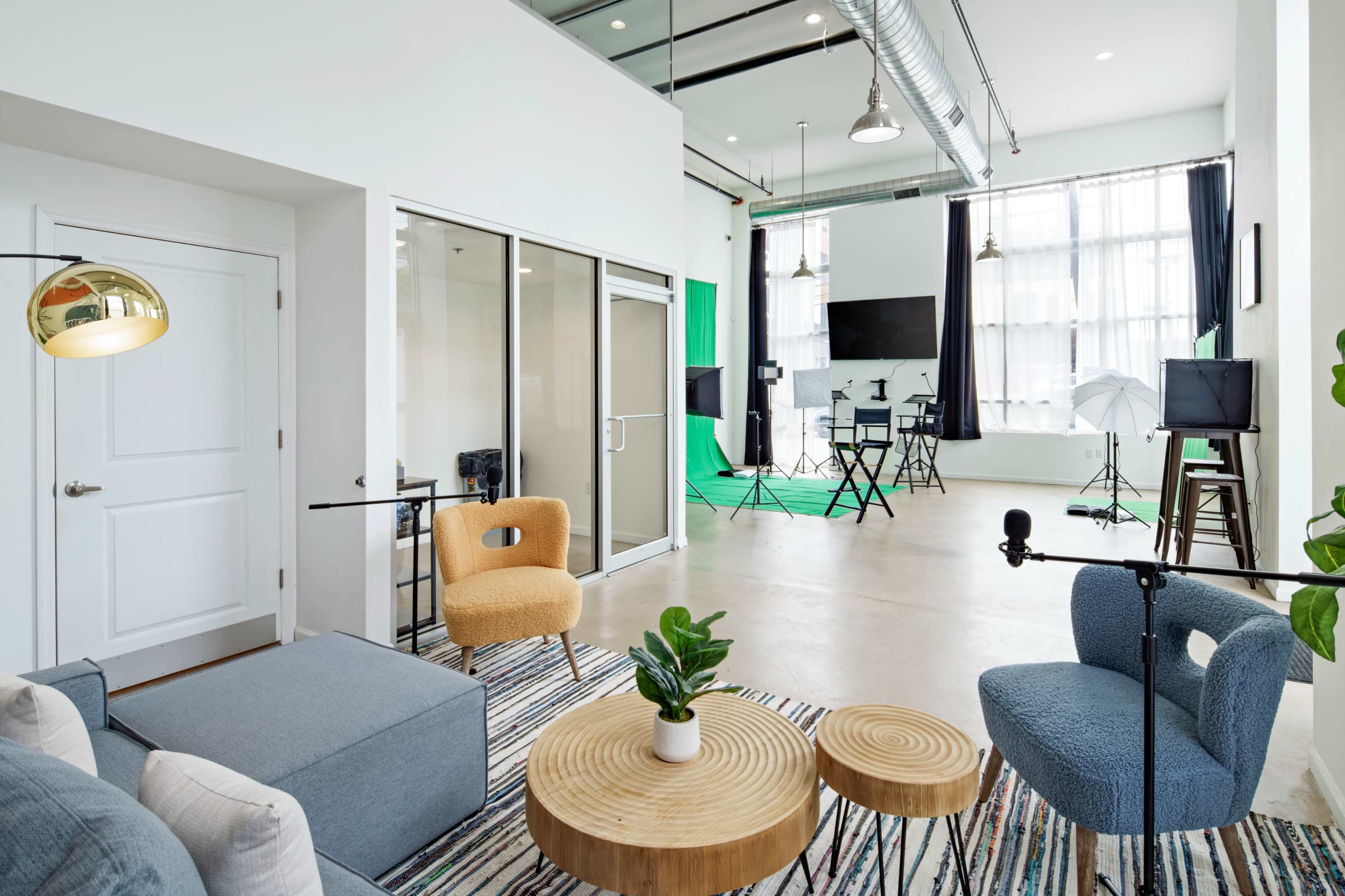 A modern studio space features a seating area with colorful chairs, a striped rug, and a green screen setup with lighting equipment in the background.