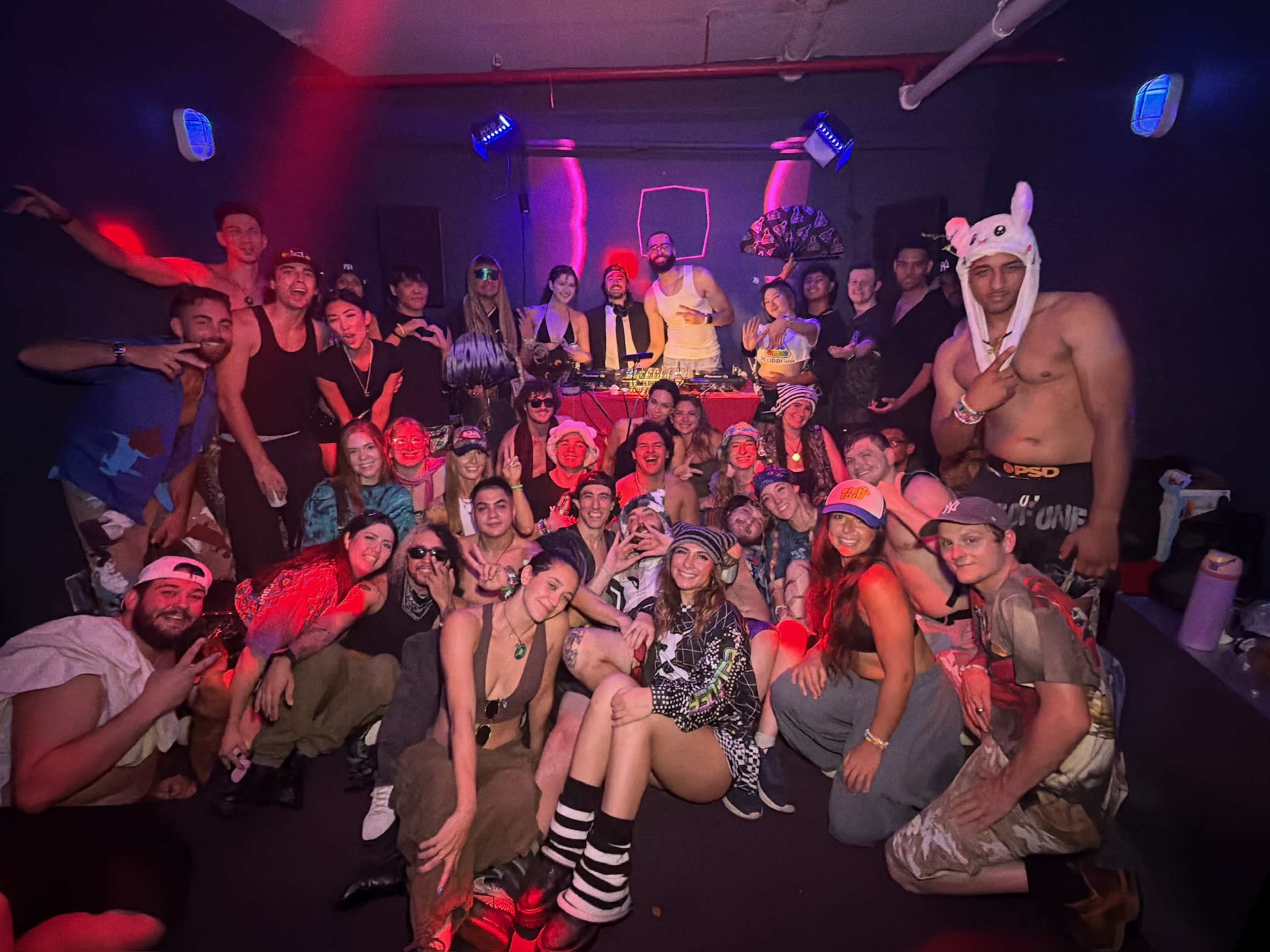 A large group of people is gathered in a club-like setting, posing for a photo in front of a DJ booth with colorful lighting.