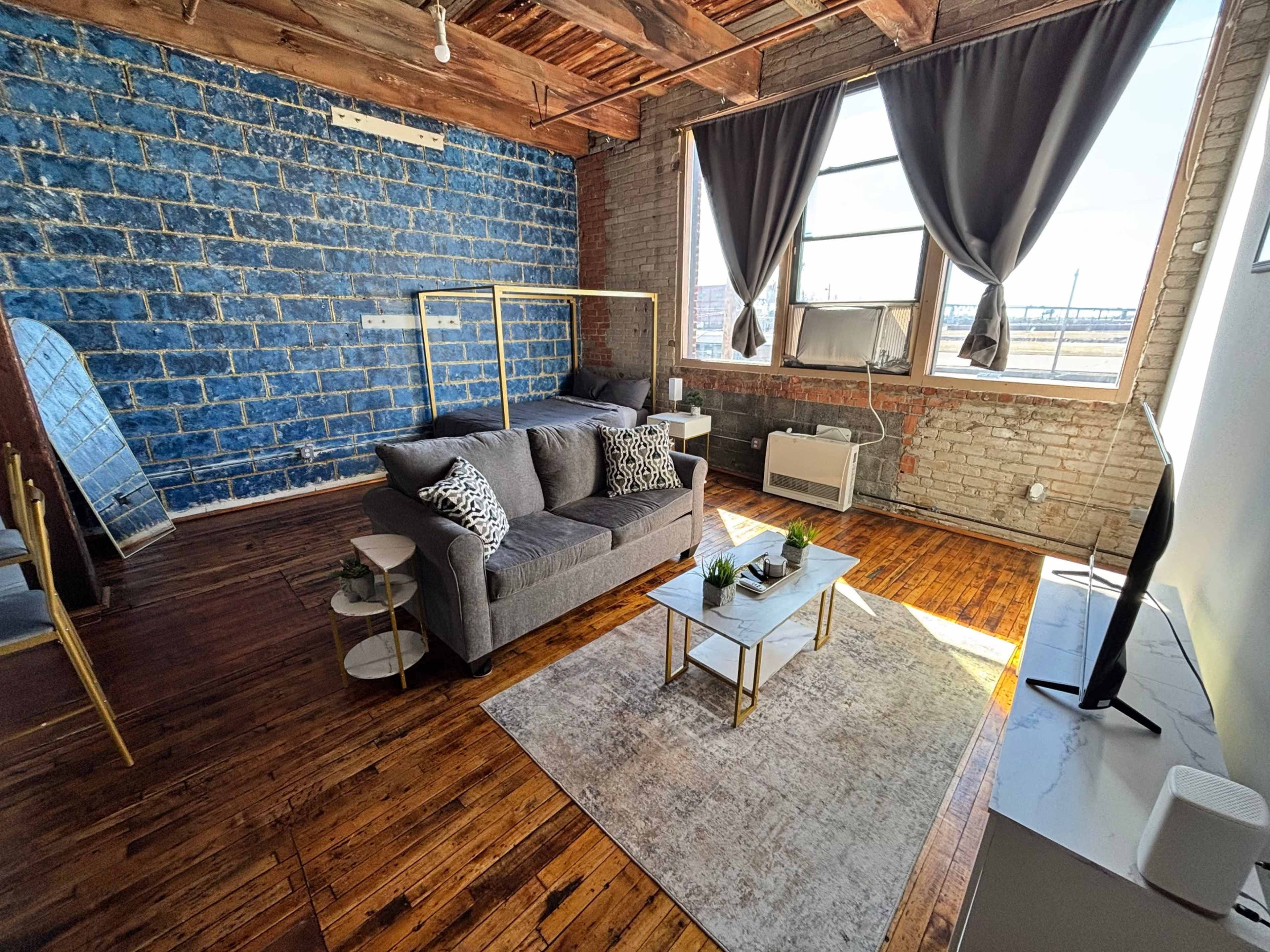 The image shows a modern living room with a blue brick wall, a gray couch with patterned pillows, a wooden floor, and large windows dressed with dark curtains.
