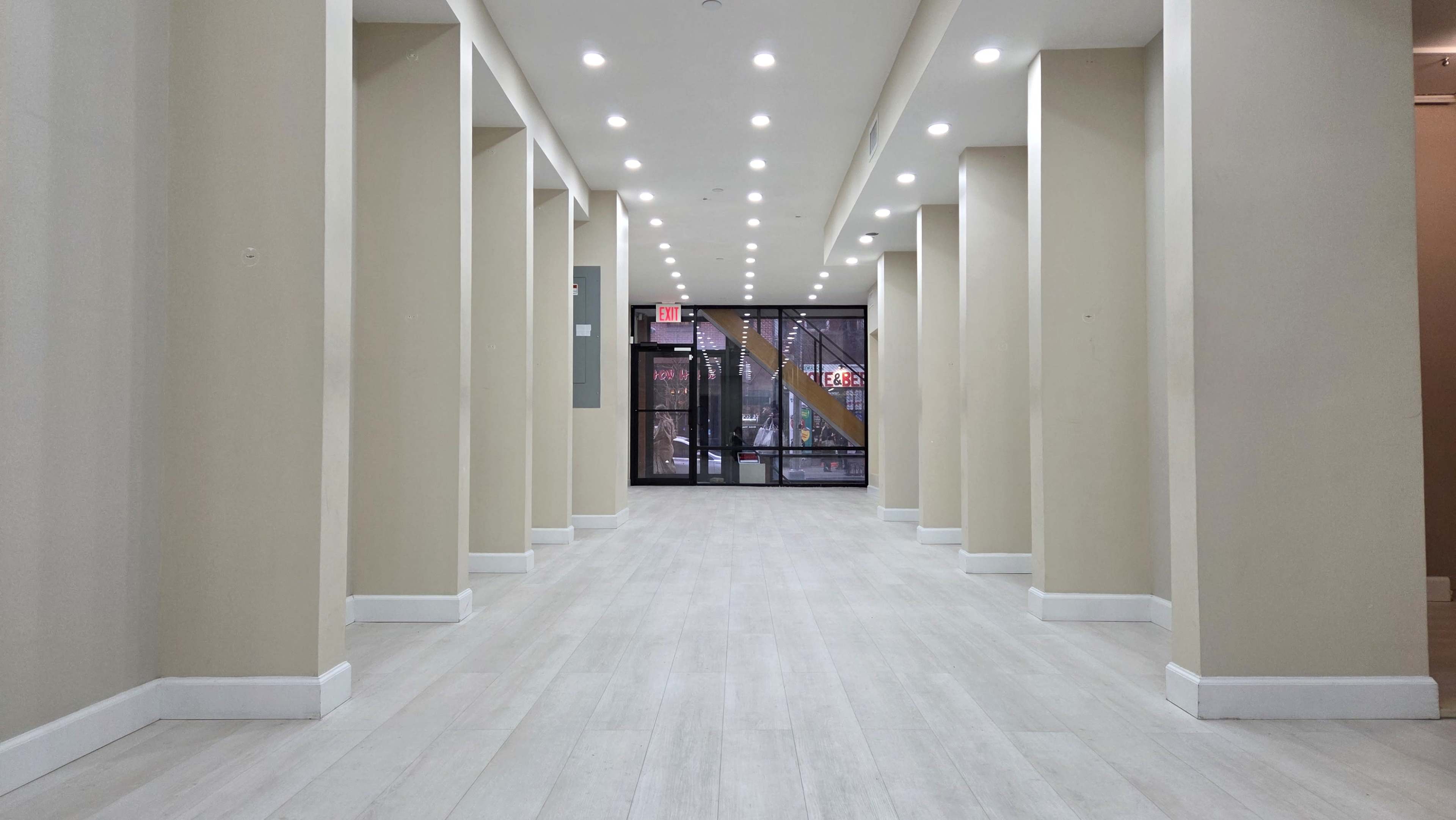 The image shows a hallway with multiple columns, lined with overhead lights, leading to a glass door entrance.