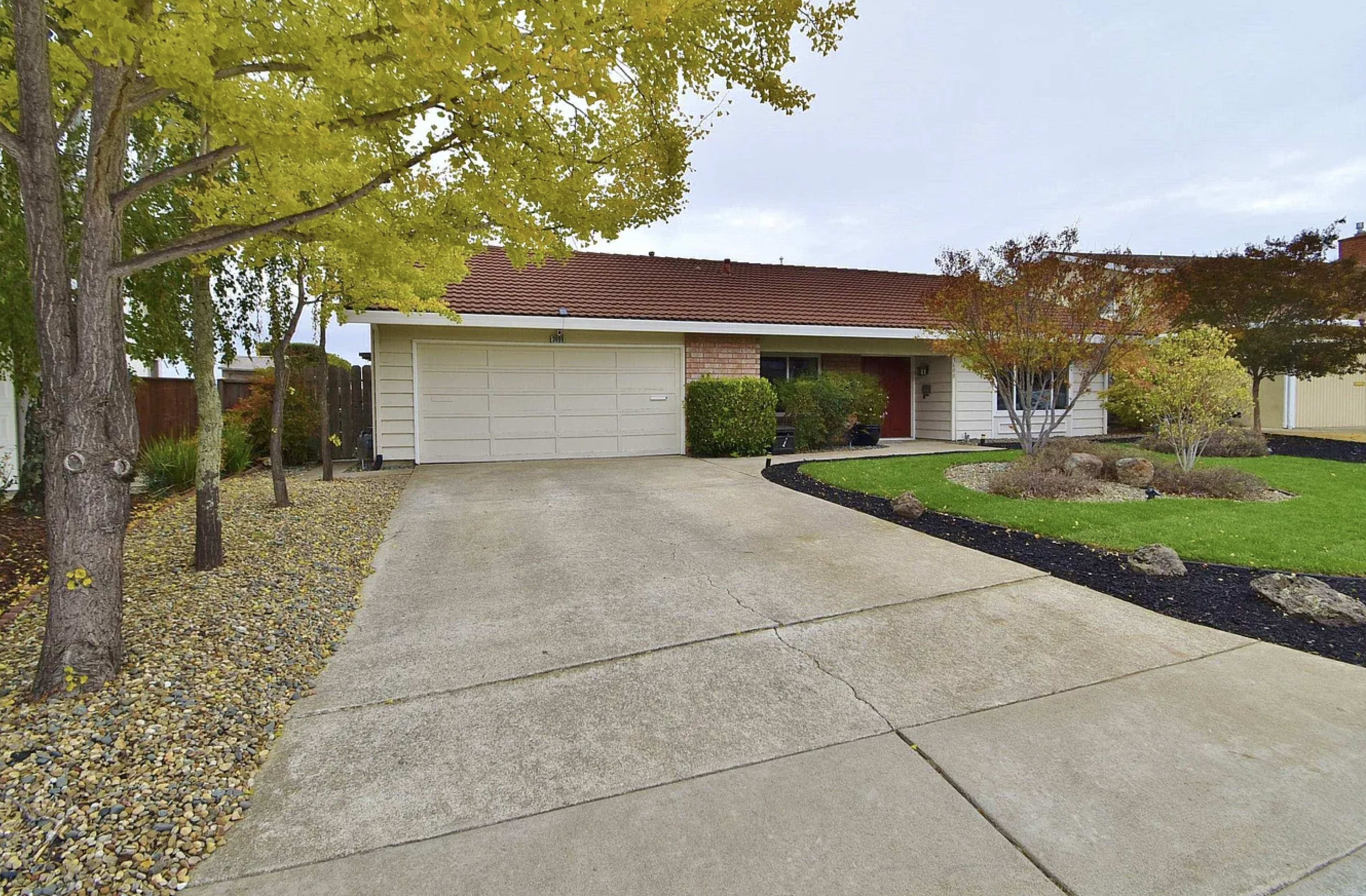 The image shows a single-story house with a garage, a paved driveway, and landscaped front yard featuring trees and plants.