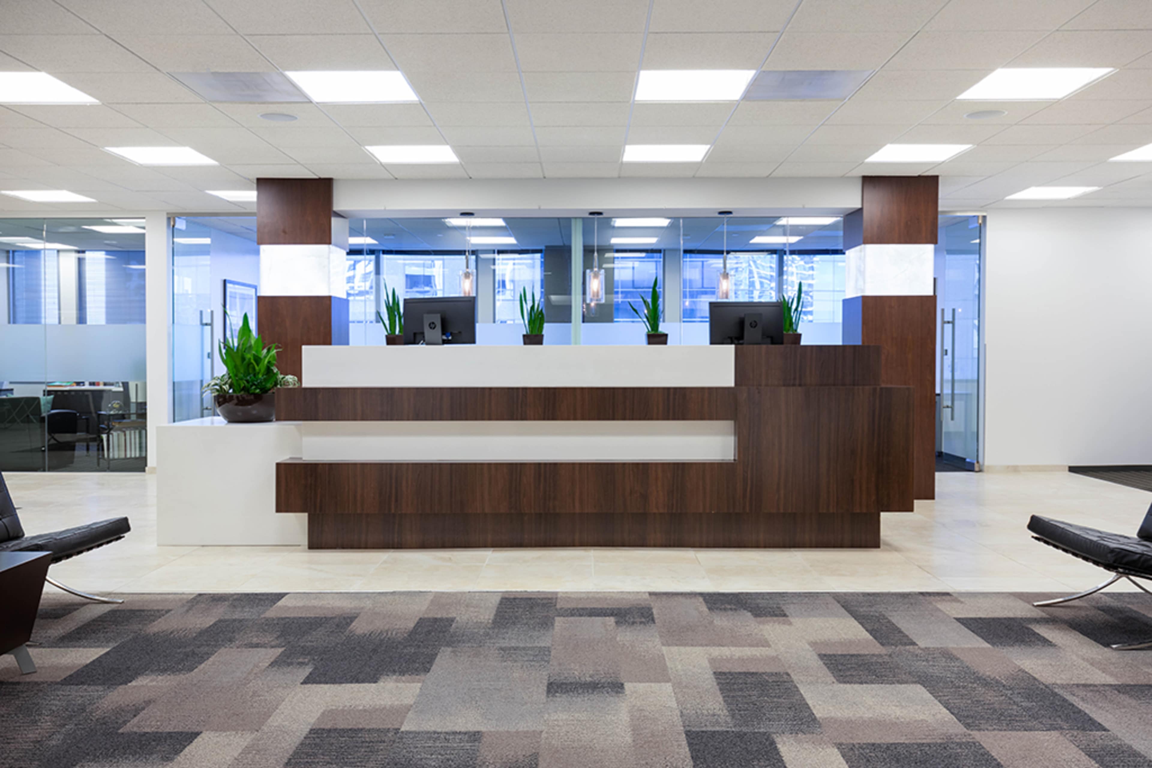 A modern reception area with a wooden front desk and two computer monitors, surrounded by glass walls and potted plants.