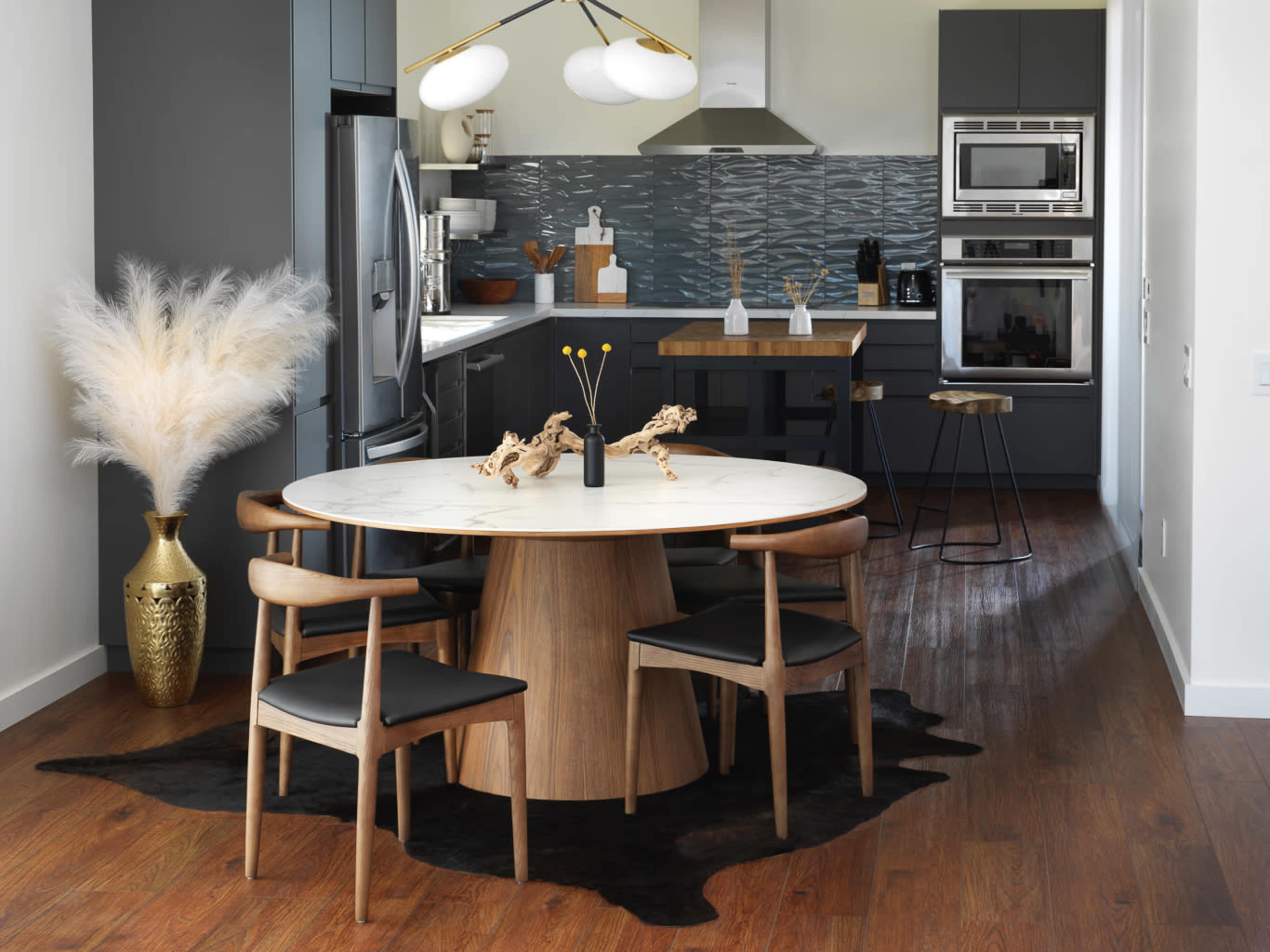 A modern dining area features a round wooden table surrounded by four black chairs, with a decorative vase and a large plant, all set against a sleek kitchen backdrop.