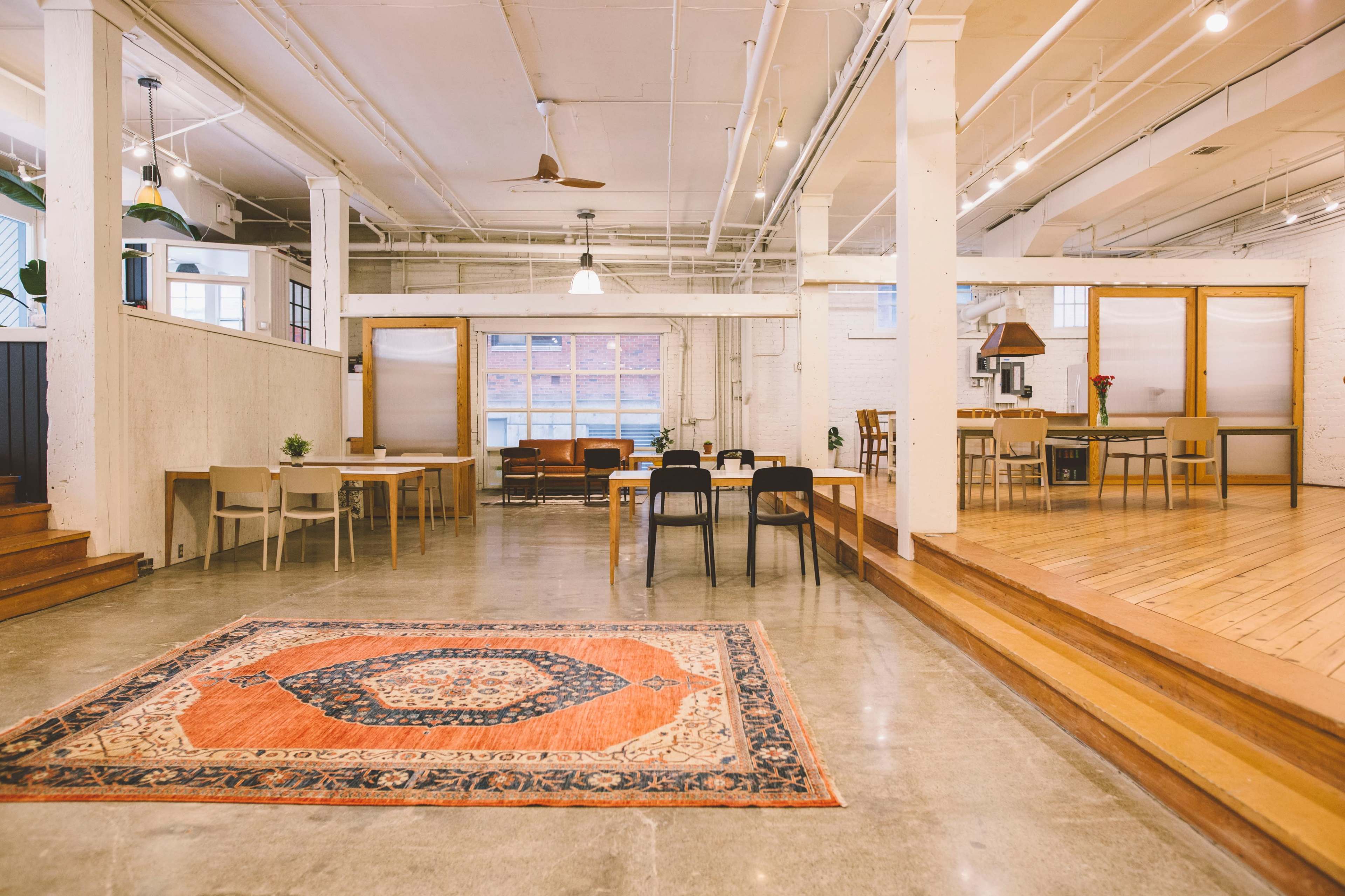 A spacious, modern interior with a patterned area rug, multiple tables and chairs, and large windows allowing natural light.