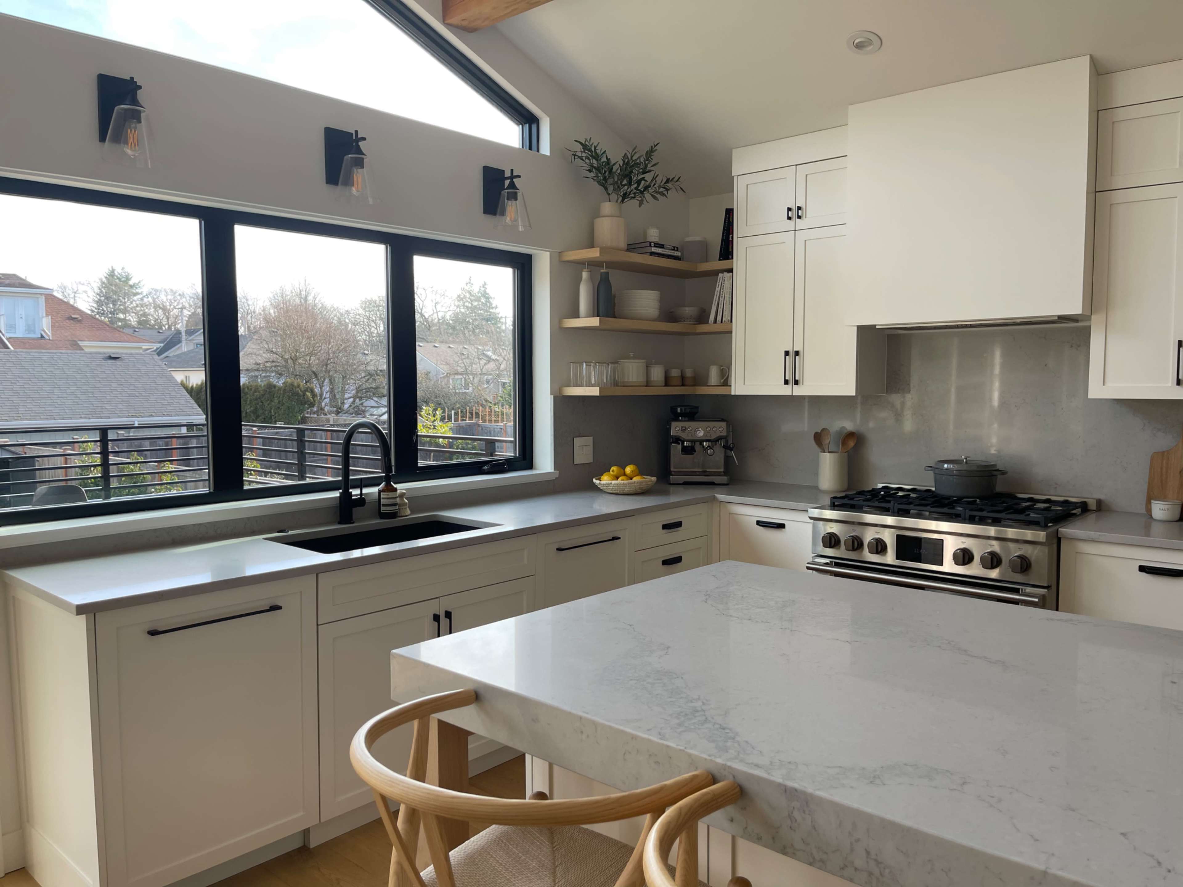 A modern kitchen features white cabinetry, a large stone countertop, and large windows overlooking an outdoor space.