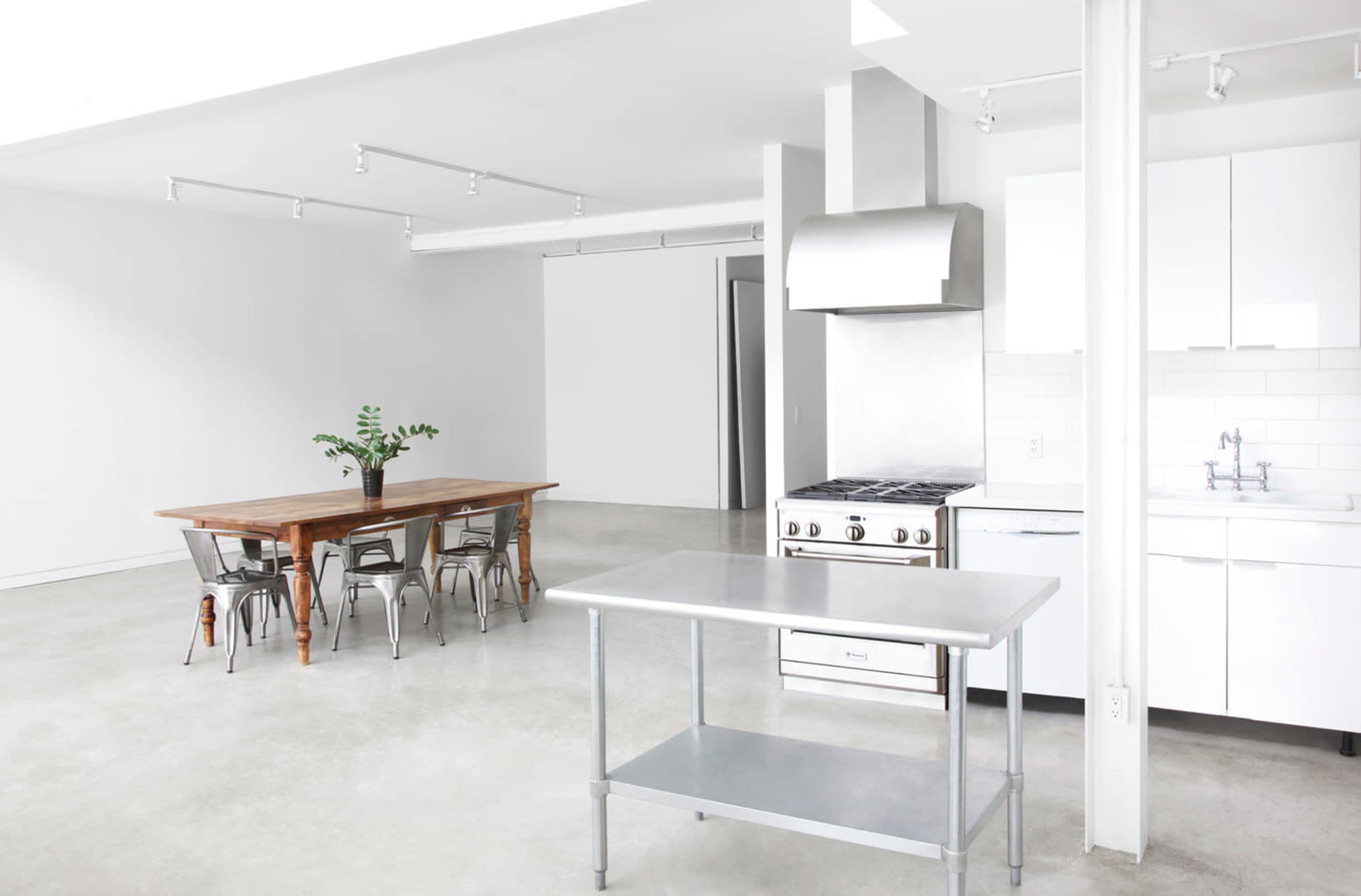 A modern kitchen features a stainless steel stove and a central prep table, with a dining area that includes a wooden table and metal chairs.