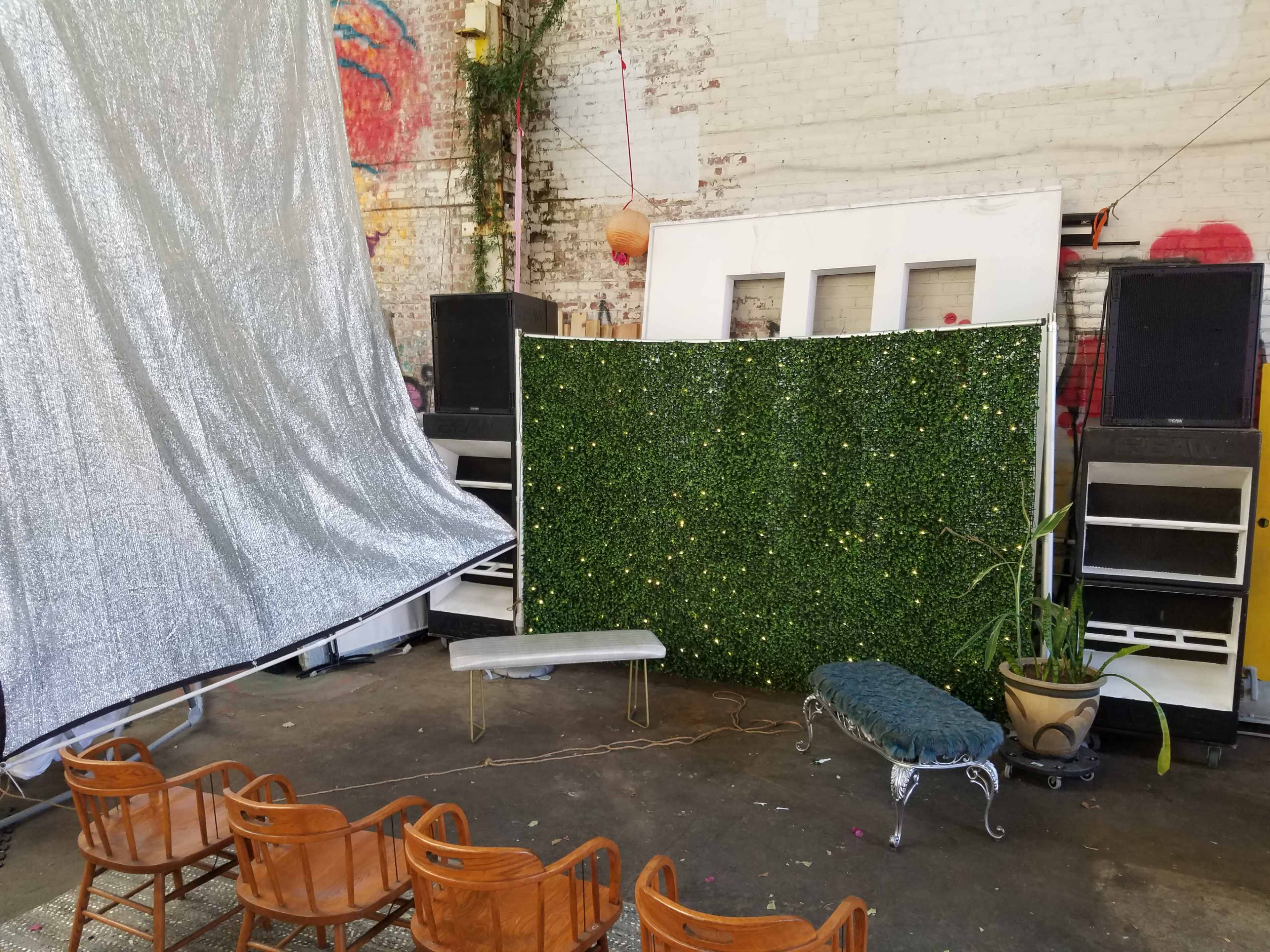 The image shows an indoor space with a green wall covered in artificial plants, a silver draped area, seating arranged in front, and large sound speakers in the background.
