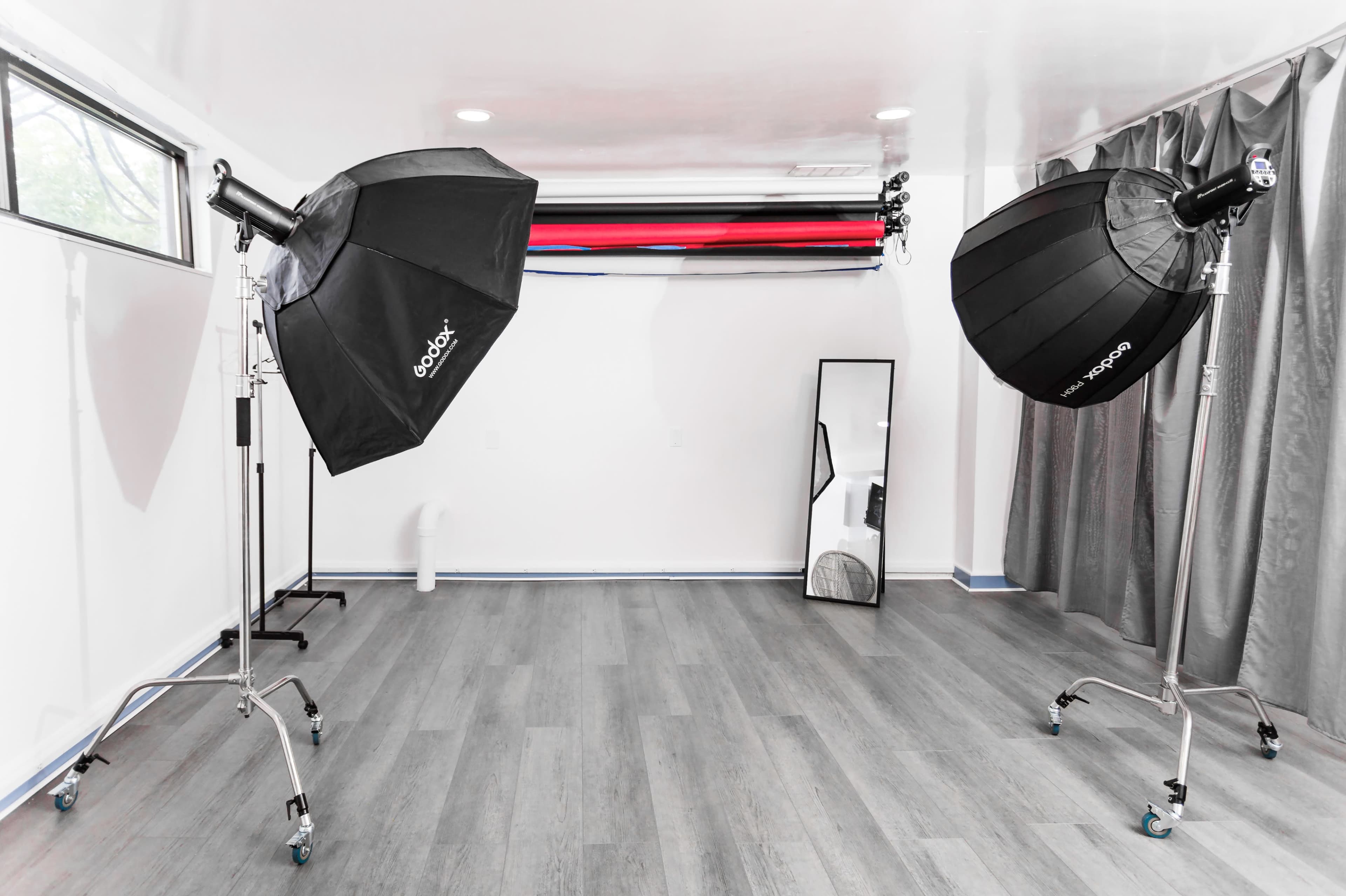 A photography studio with two light softboxes on stands, a large mirror, and a backdrop setup featuring red and black rolls.