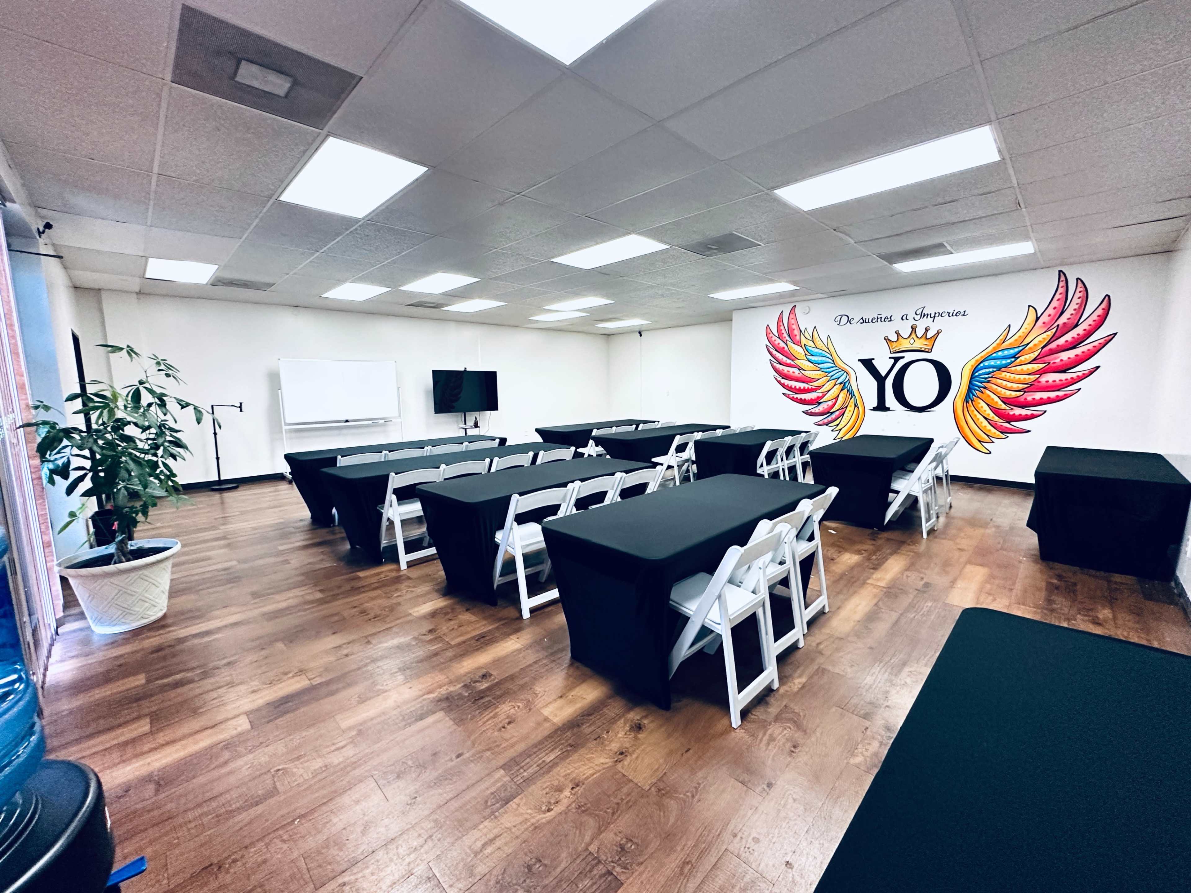 The image shows a spacious meeting room with multiple tables covered in black cloth, arranged in a U-shape, and featuring a large mural with the word "YO" adorned with colorful wings on one wall.