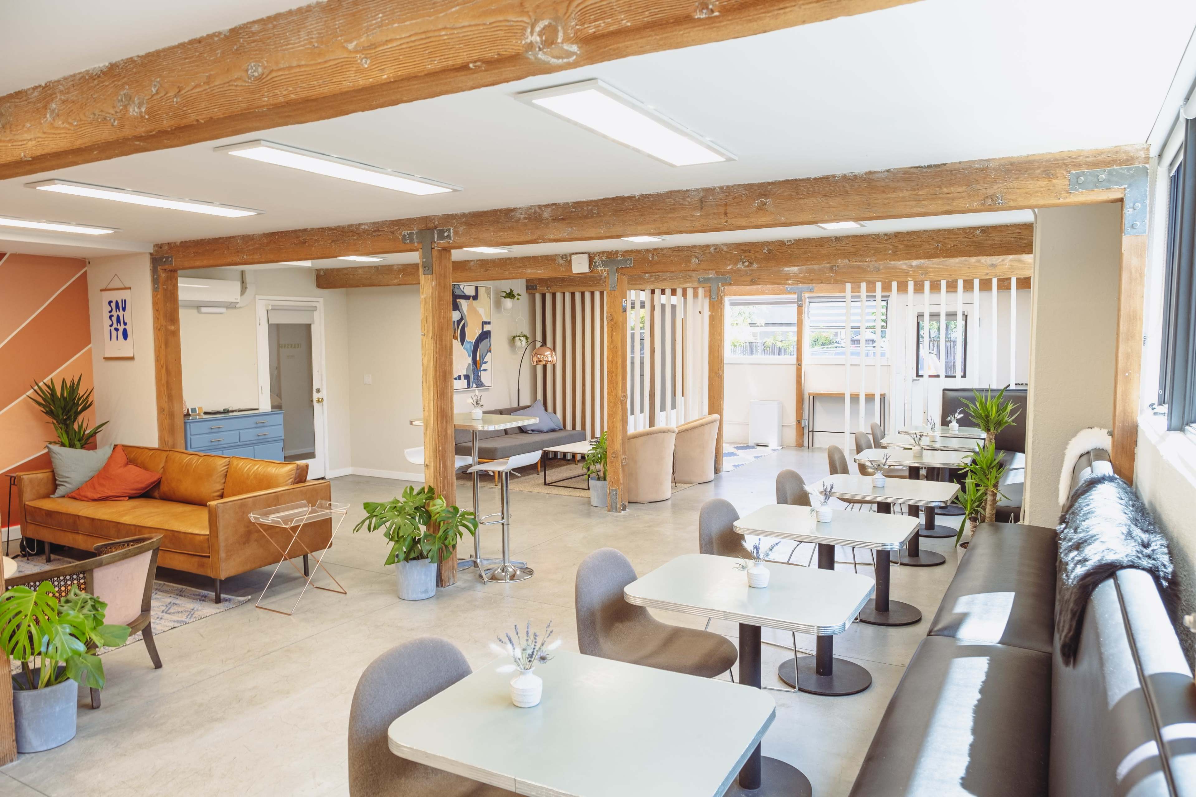 A spacious interior features a mix of seating areas, including sofas and tables, surrounded by plants and wooden beams.