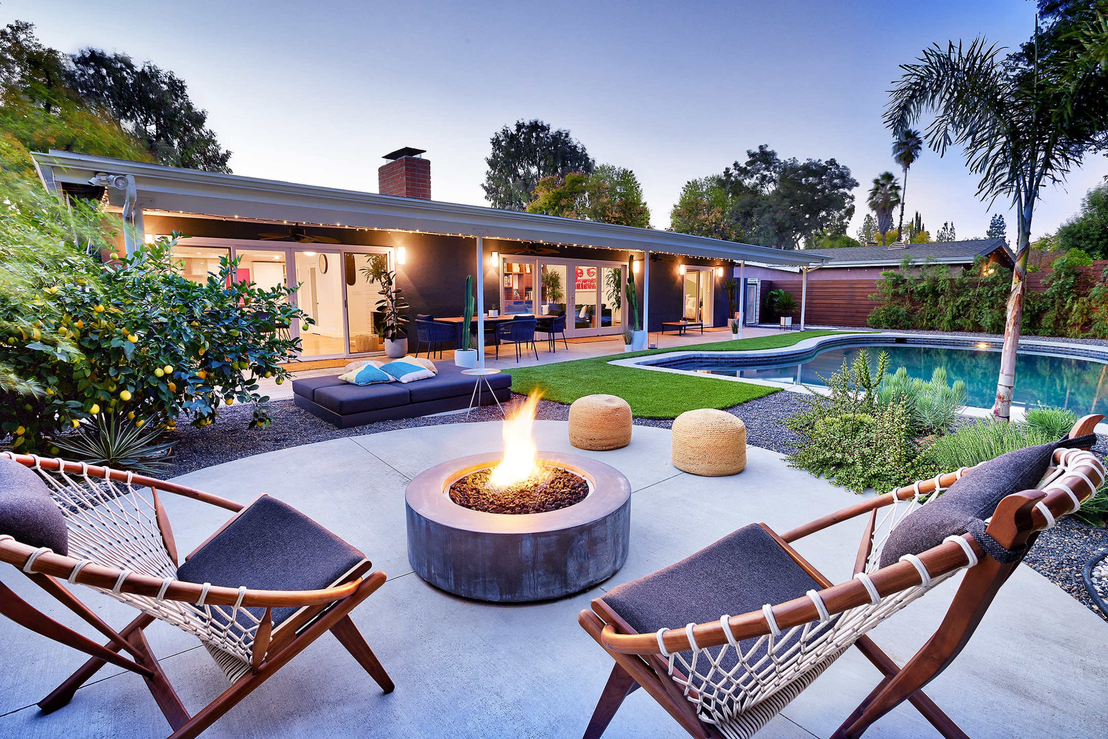 A modern backyard features a fire pit surrounded by two chairs, an inviting swimming pool, and a well-kept lawn, with a home visible in the background.