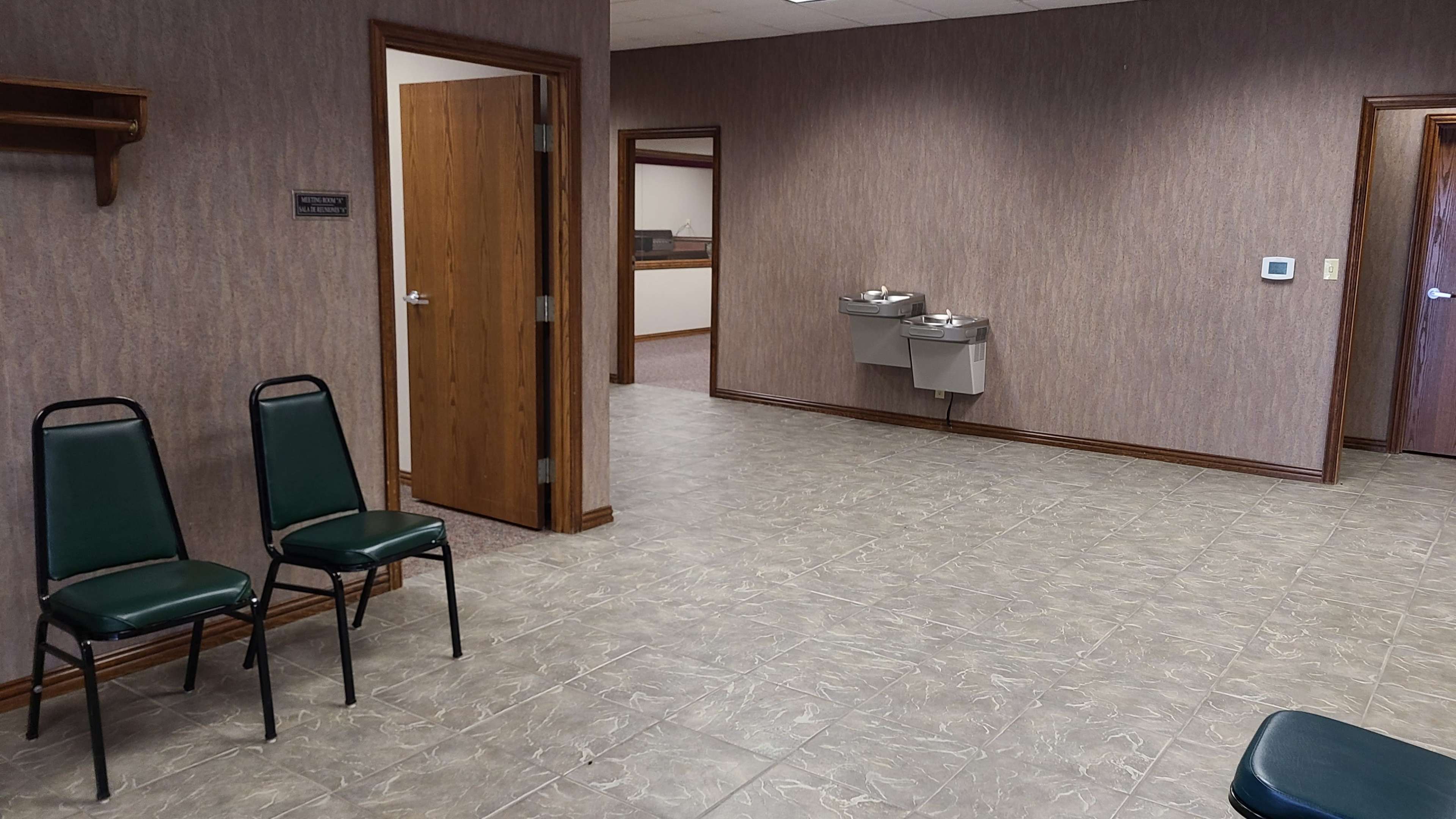 The image shows a waiting area with two green chairs, a closed door, and two drinking fountains on the wall opposite.