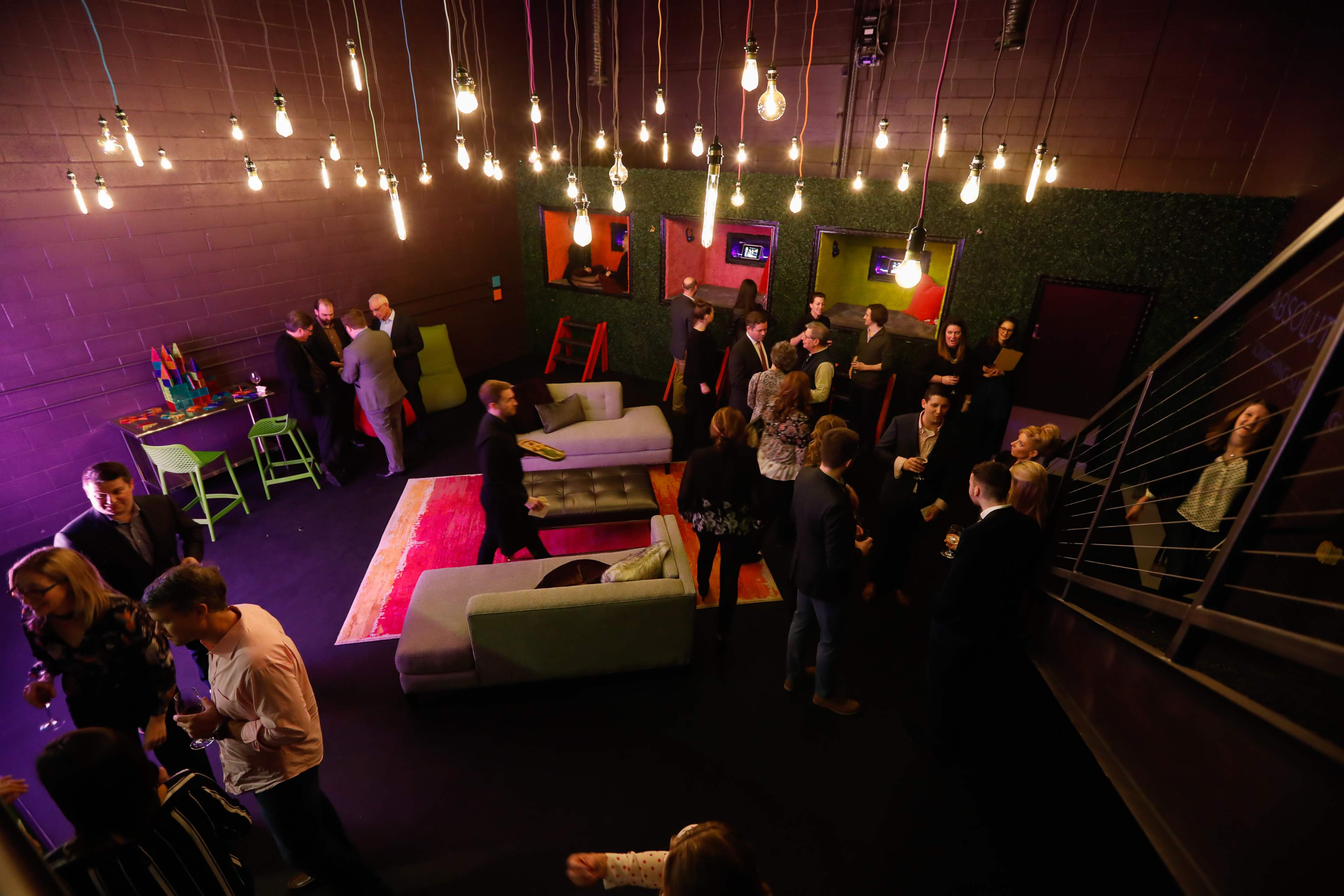 A group of people socialize in a dimly lit lounge adorned with green plants, colorful artwork, and hanging light bulbs.